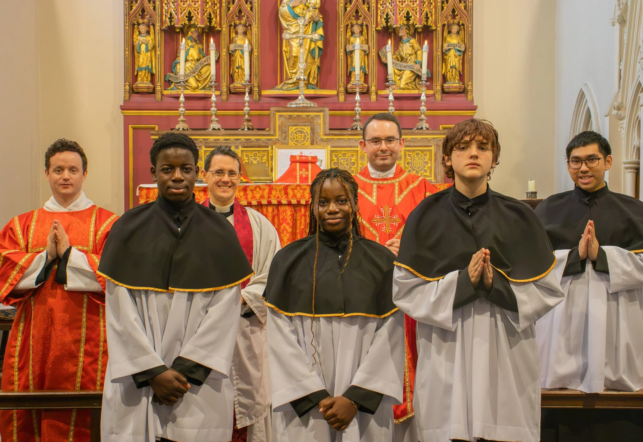 Group of seven people dressed in religious robes standing in front of an ornate church altar with candles and religious statues.