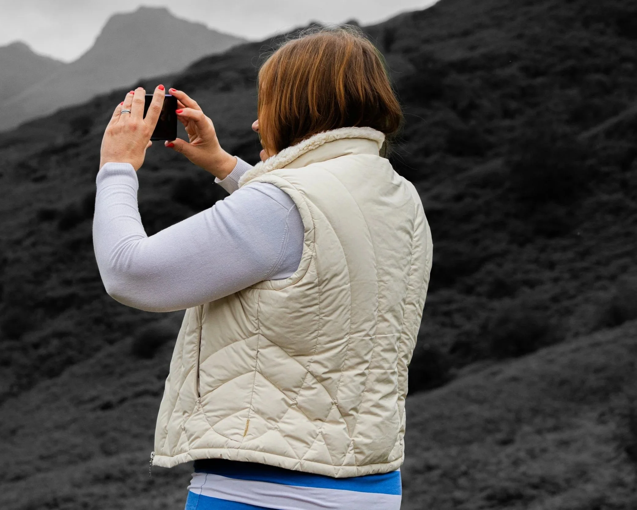 A woman with reddish-brown hair taking a photo with her smartphone in an outdoor setting with dark, volcanic terrain and mountains in the background.