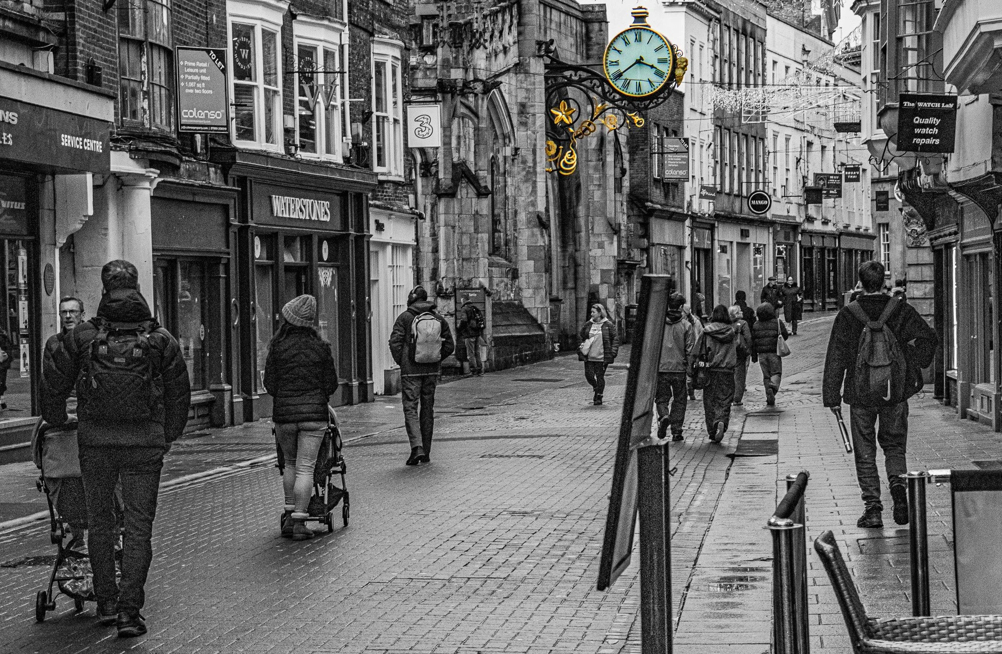A black and white street scene with people walking and a large blue clock with gold details hanging above. The street has shops on both sides and is wet, possibly after rain.