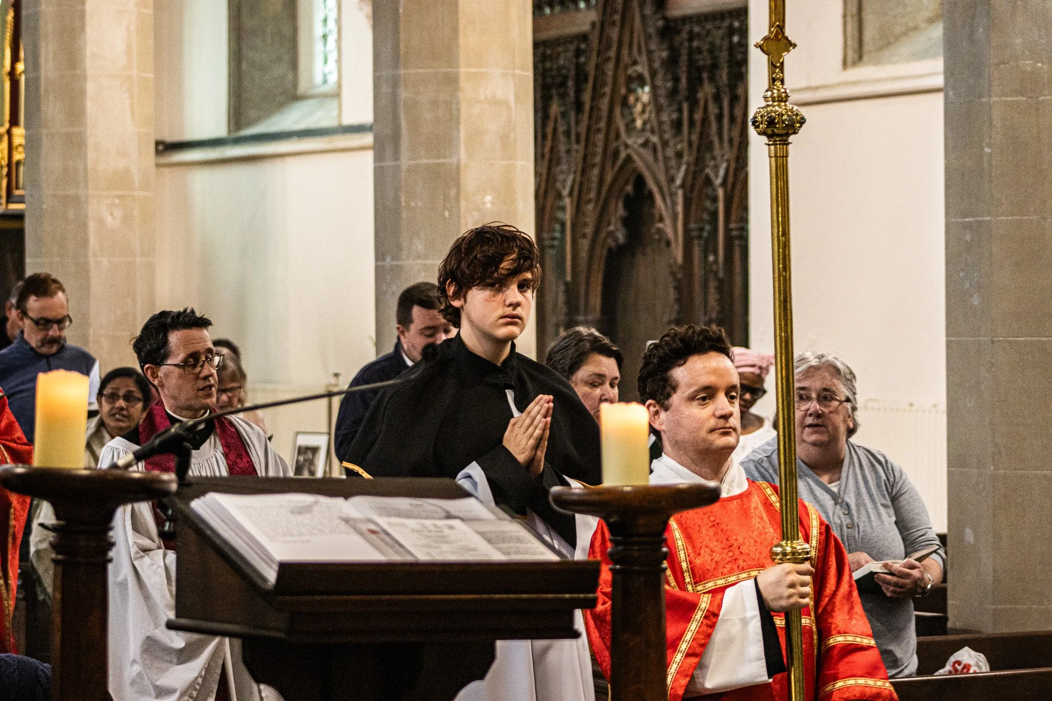 A group of people participating in a religious ceremony inside a church, with a young man at the center praying, others praying, and clergy members present. The church features stained glass windows and ornate wooden decorations.
