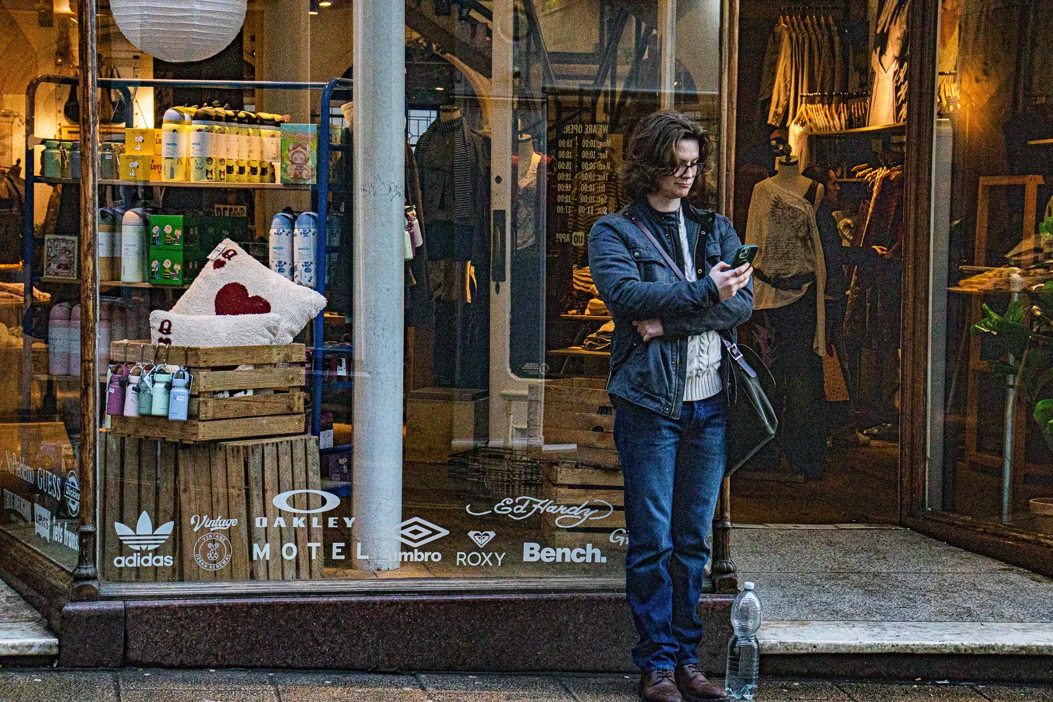 A woman with curly hair and glasses stands outside a store, looking at her phone. The store window behind her displays clothing and accessories, and the window has various brand logos at the bottom.