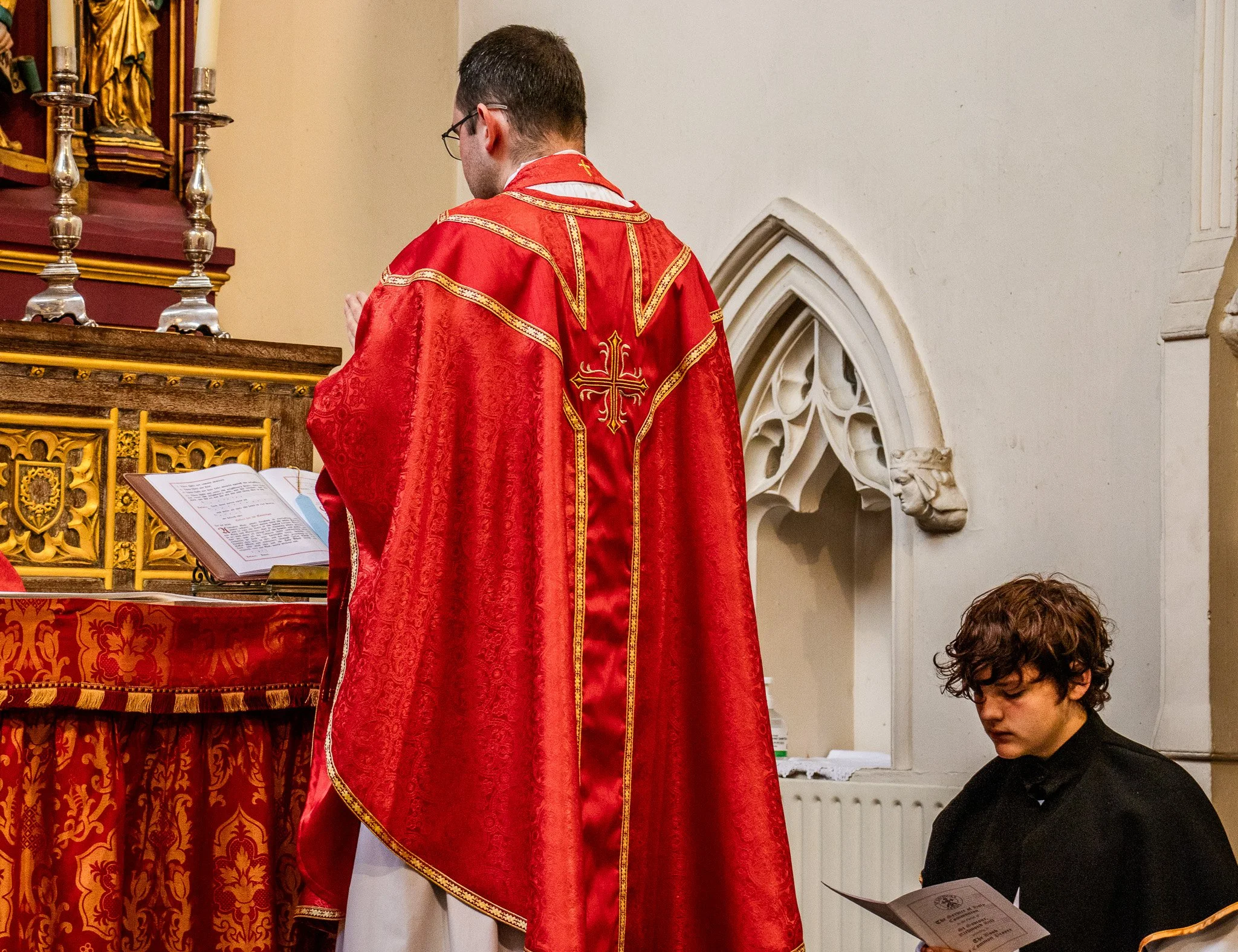 A priest in red robes with gold embroidery praying at an altar inside a church.