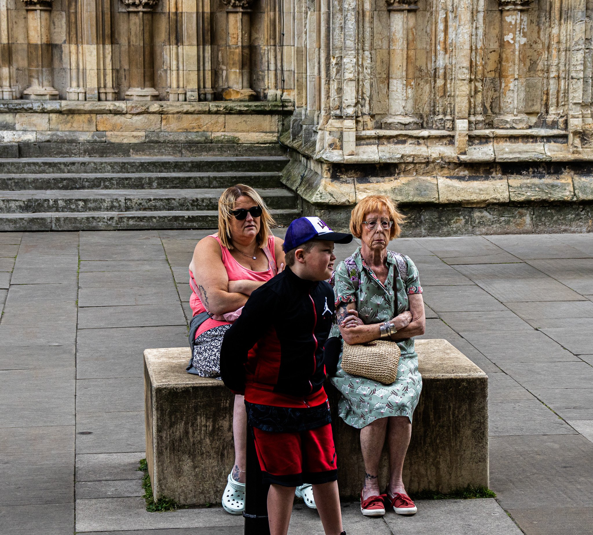 Four people sitting on steps in front of a stone building with columns. Two women in the foreground, one wearing sunglasses and a pink top, the other older with glasses and a floral shirt. A boy with a cap and a woman with sunglasses are also visible