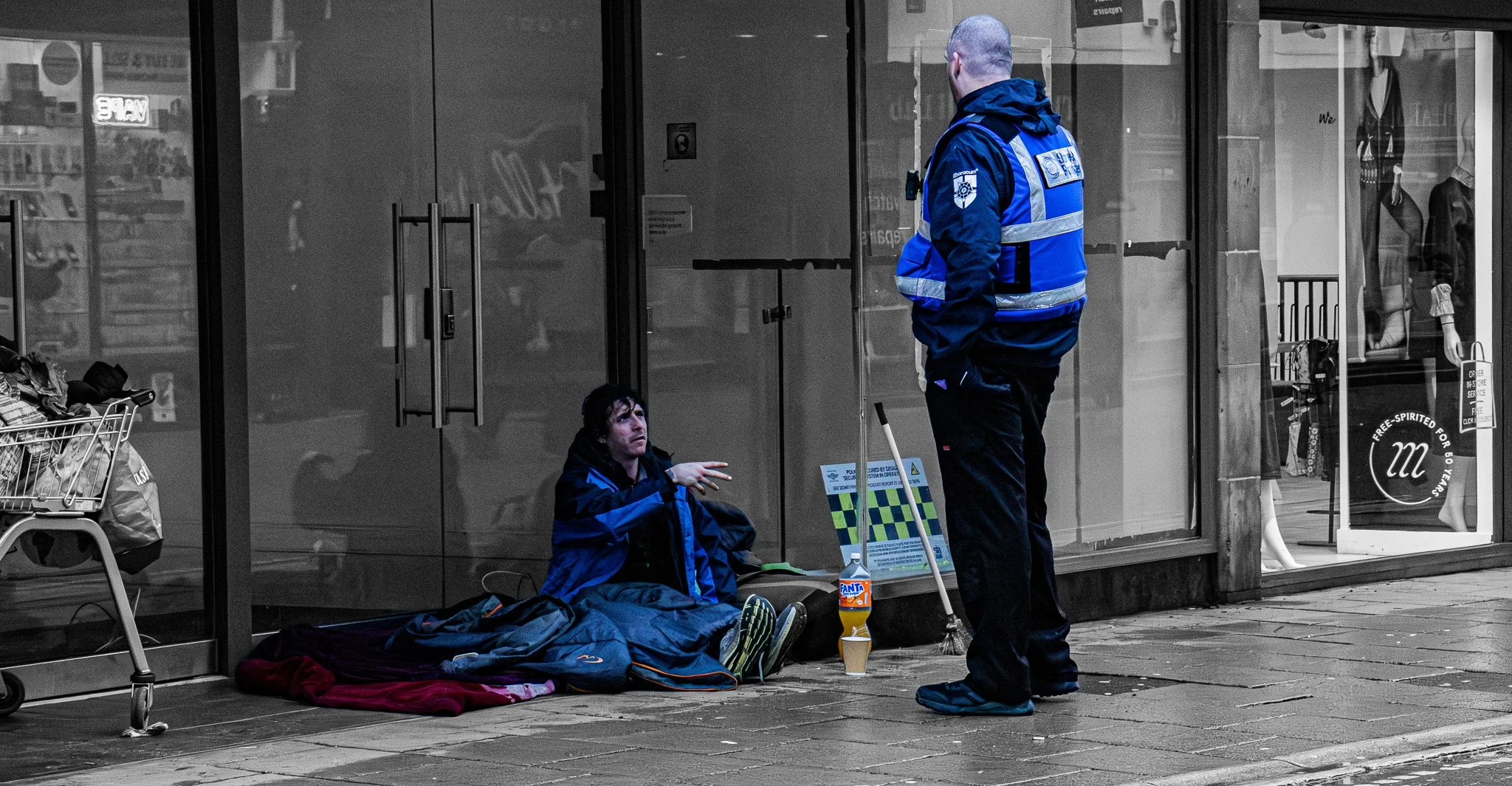Two men are sitting and standing outside a storefront, dressed in blue winter jackets. One man is sitting on the ground leaning against the glass, looking up at the other man who is standing. There are bags, a skateboard, and a bottle of Fanta nearby