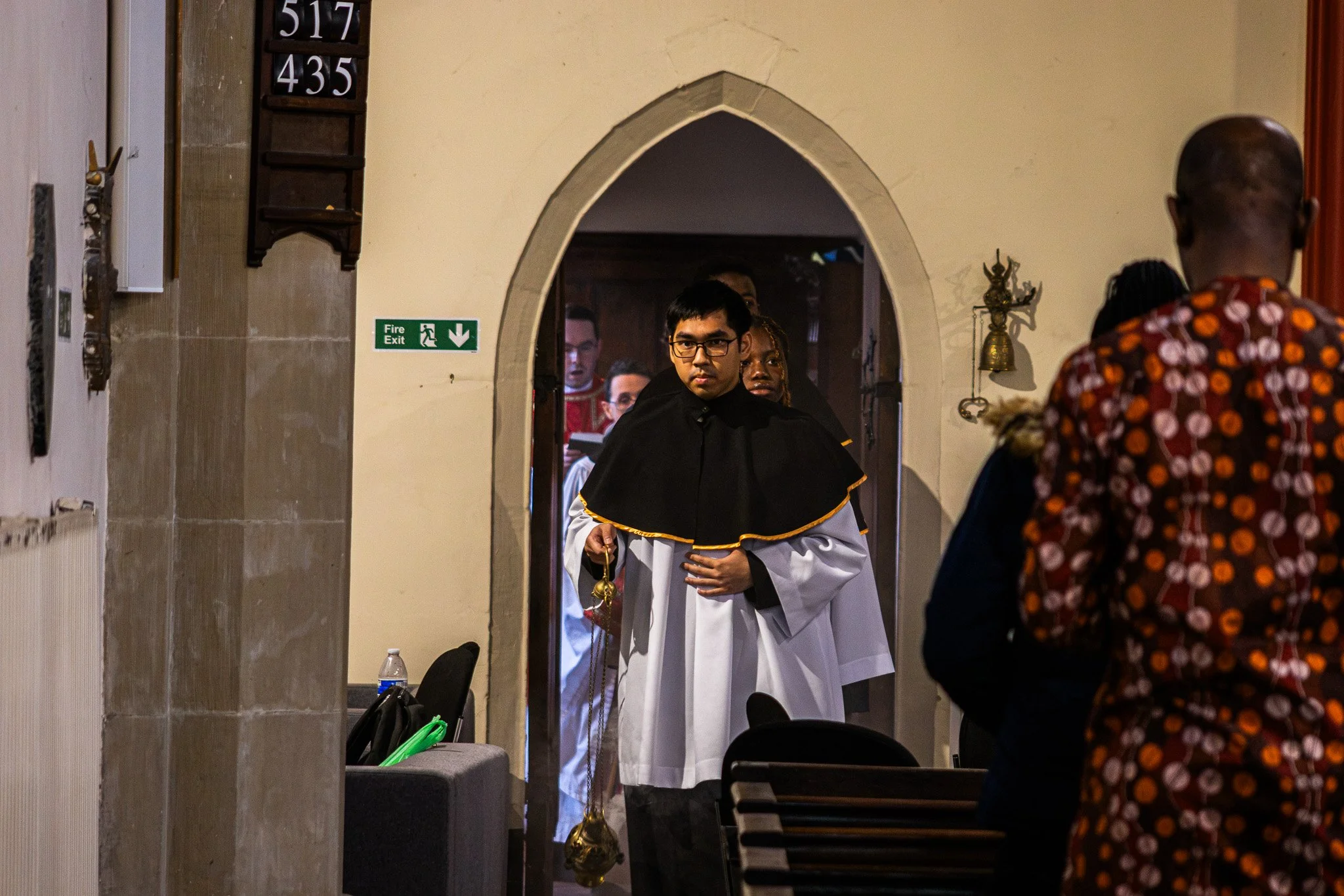 Church choir member in a white and black robe standing in a church doorway during a service, facing the camera with a serious expression, with other choir members visible behind him.