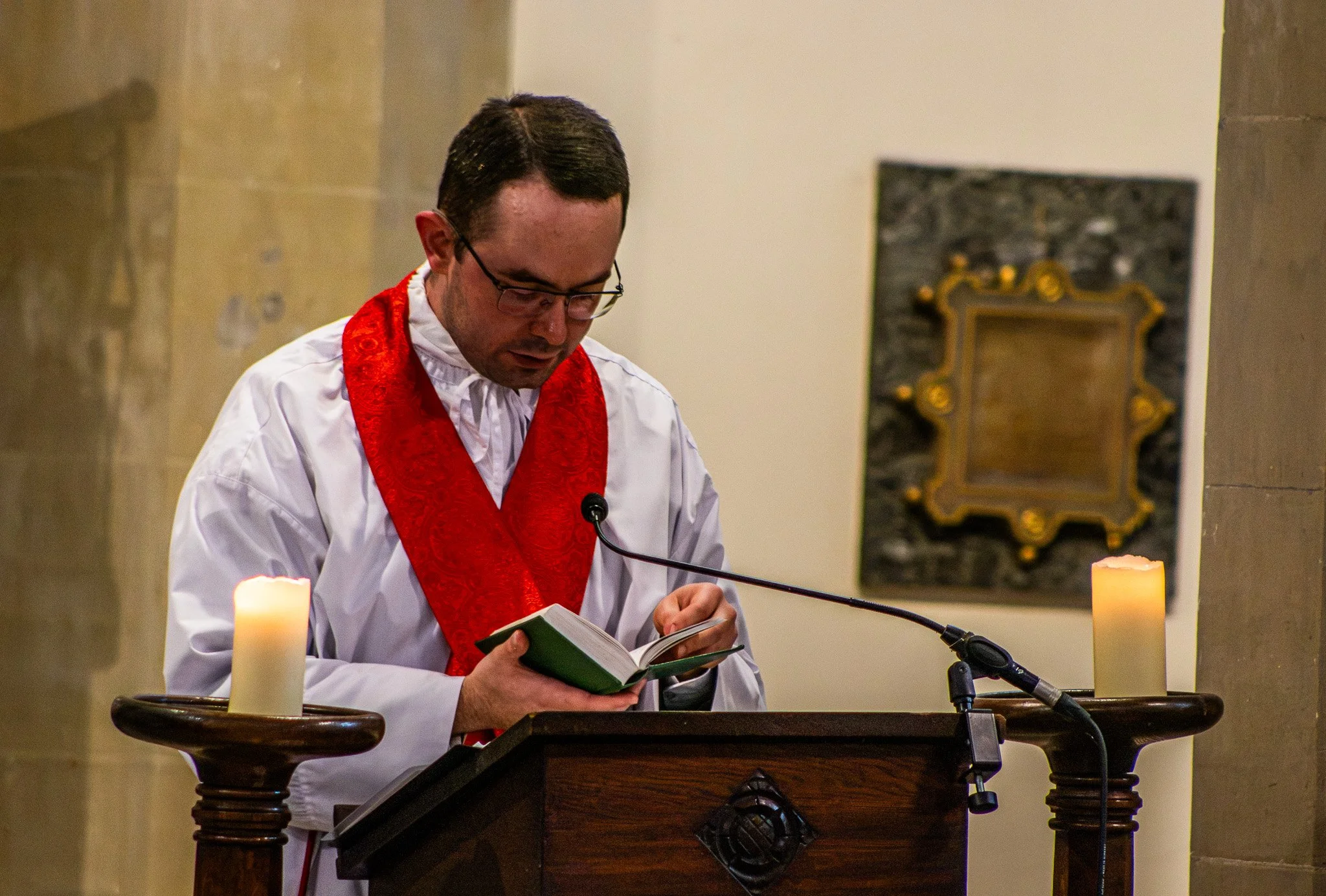 A man wearing glasses, a white robe, and a red stole is reading from a book at a church altar during a religious ceremony, with candles and a religious portrait in the background.