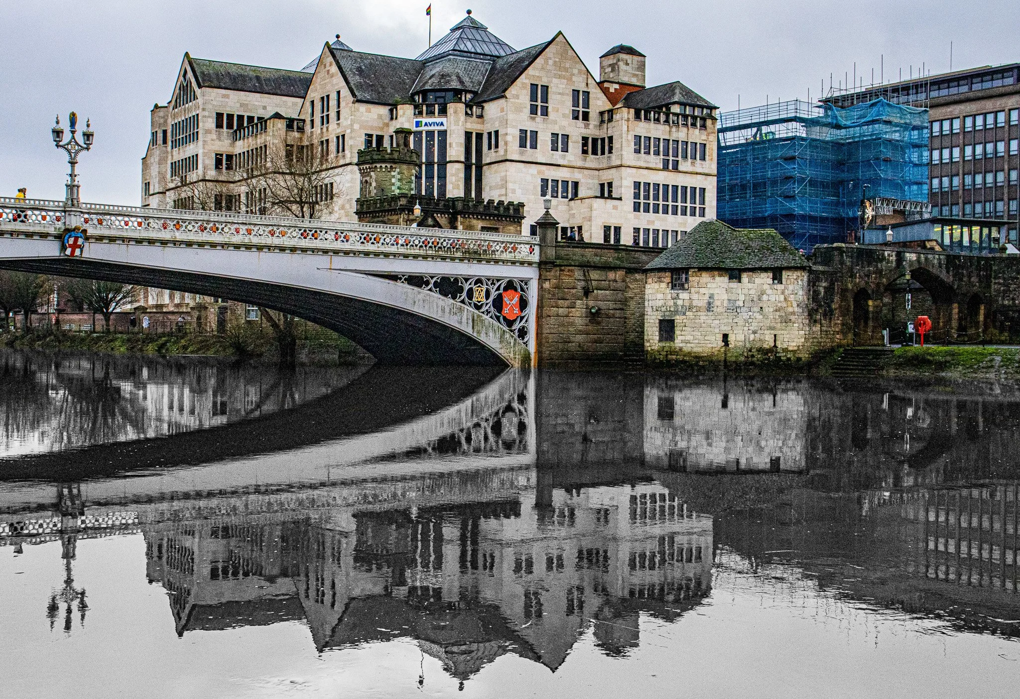 A cityscape with a bridge crossing over a river, buildings in the background, some under construction with scaffolding, and reflections of the buildings and bridge in the water.