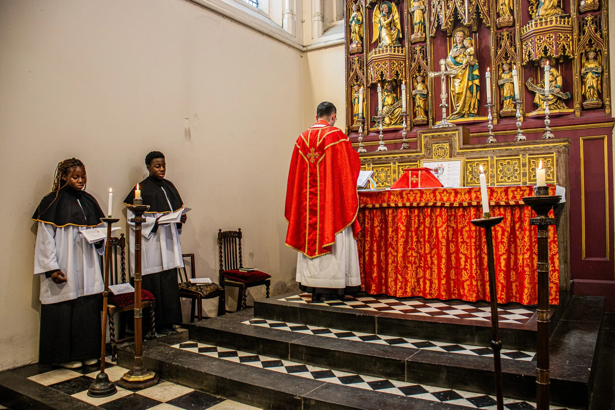 A priest and two acolytes standing before an ornate altar inside a church, with candles and religious statues.