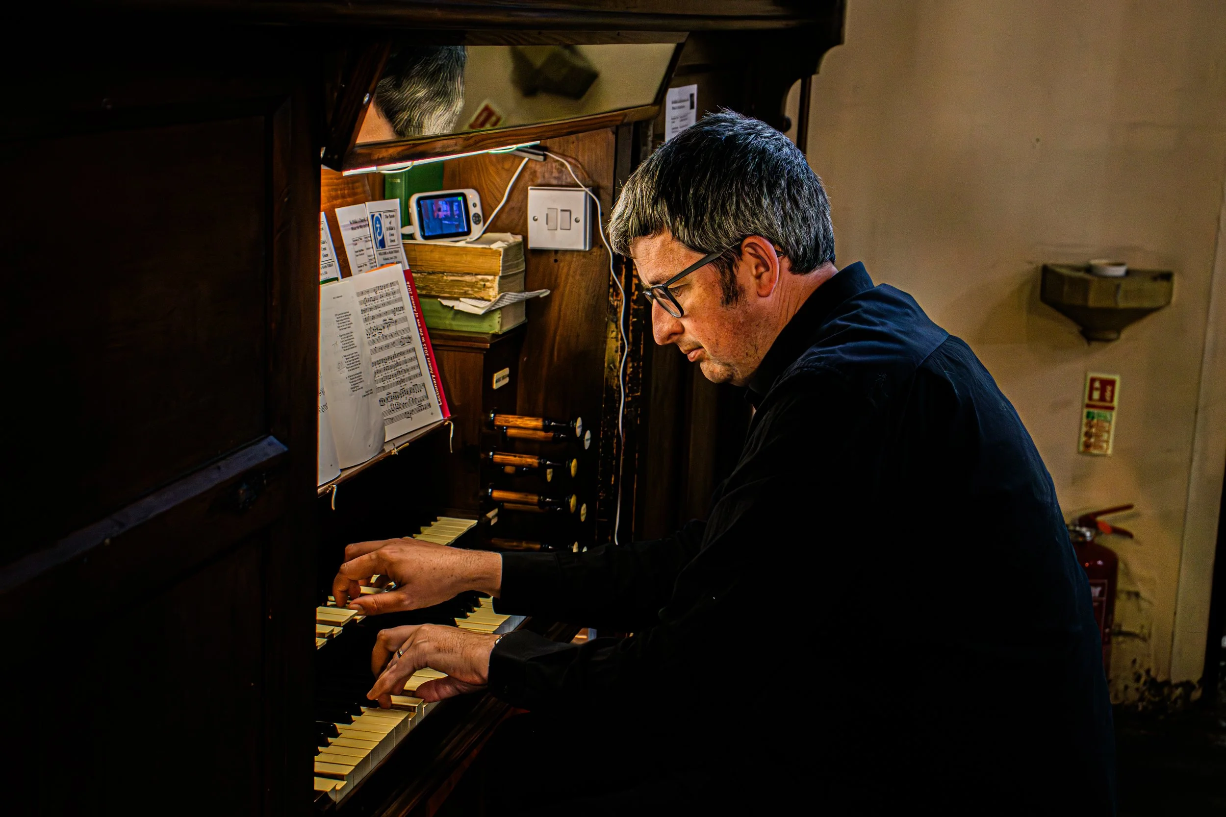 A person with glasses and dark hair playing a piano at a keyboard. The setting appears to be a cozy indoor space with books, papers, and electronic devices on the piano.