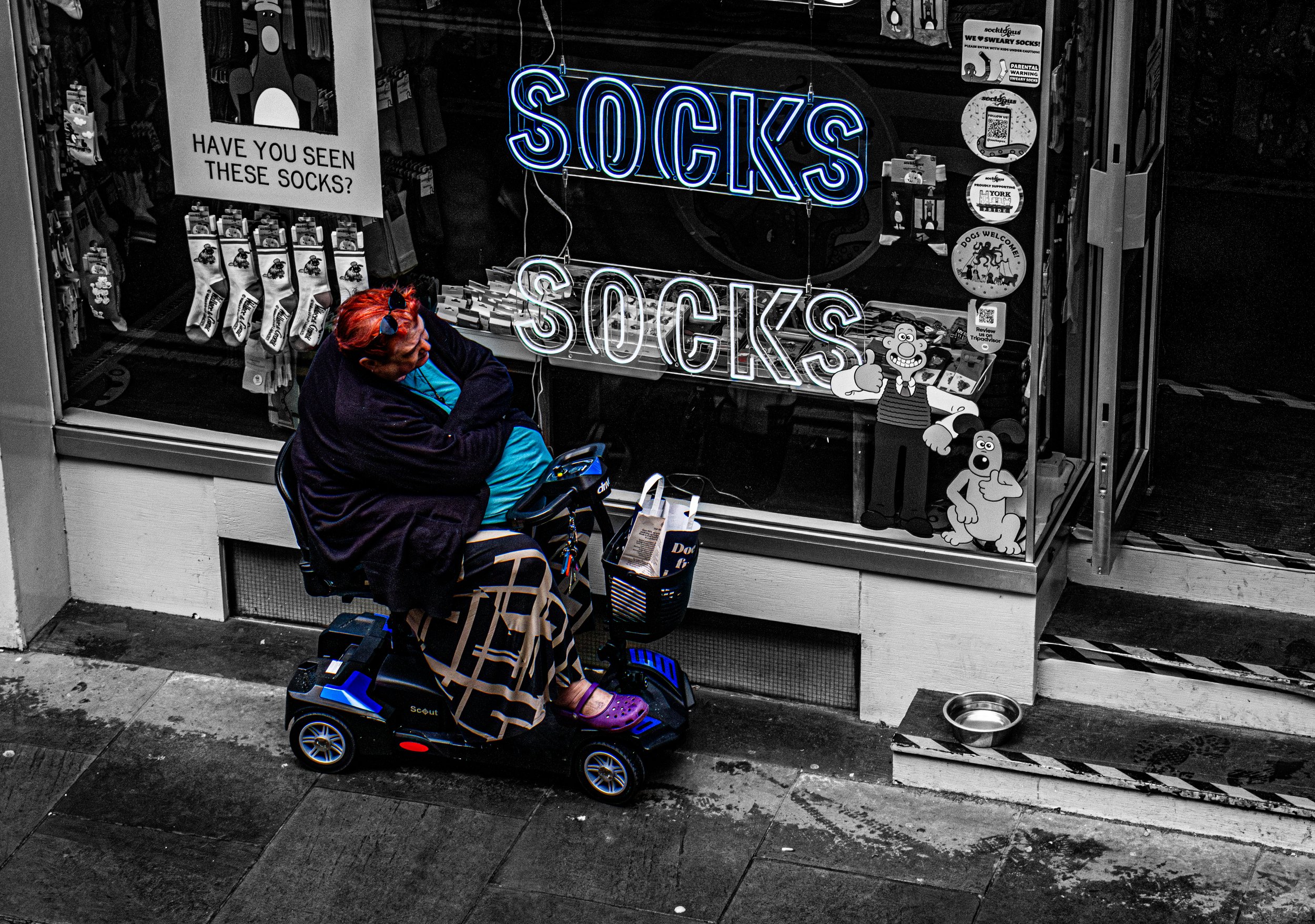 A person with red hair sitting outside a storefront with a neon sign that says 'SOCKS'. The person is wearing a dark jacket, a light blue shirt, and purple Crocs, sitting on a motorized toy car. The storefront window displays various sock packages, a