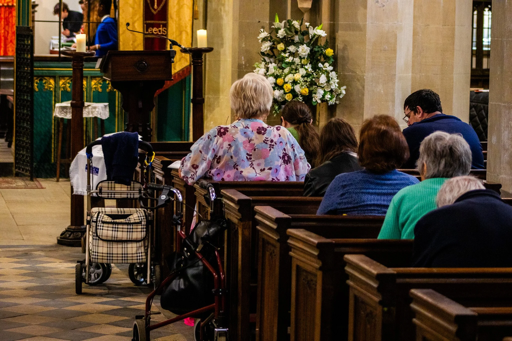 People seated inside a church or chapel, facing the altar. Some are elderly and others are younger, with various hair colors and styles. A floral arrangement with white and yellow flowers is visible. There are wheelchairs and walkers in front of the benches. The background features candles, an altar, and decorative banners.