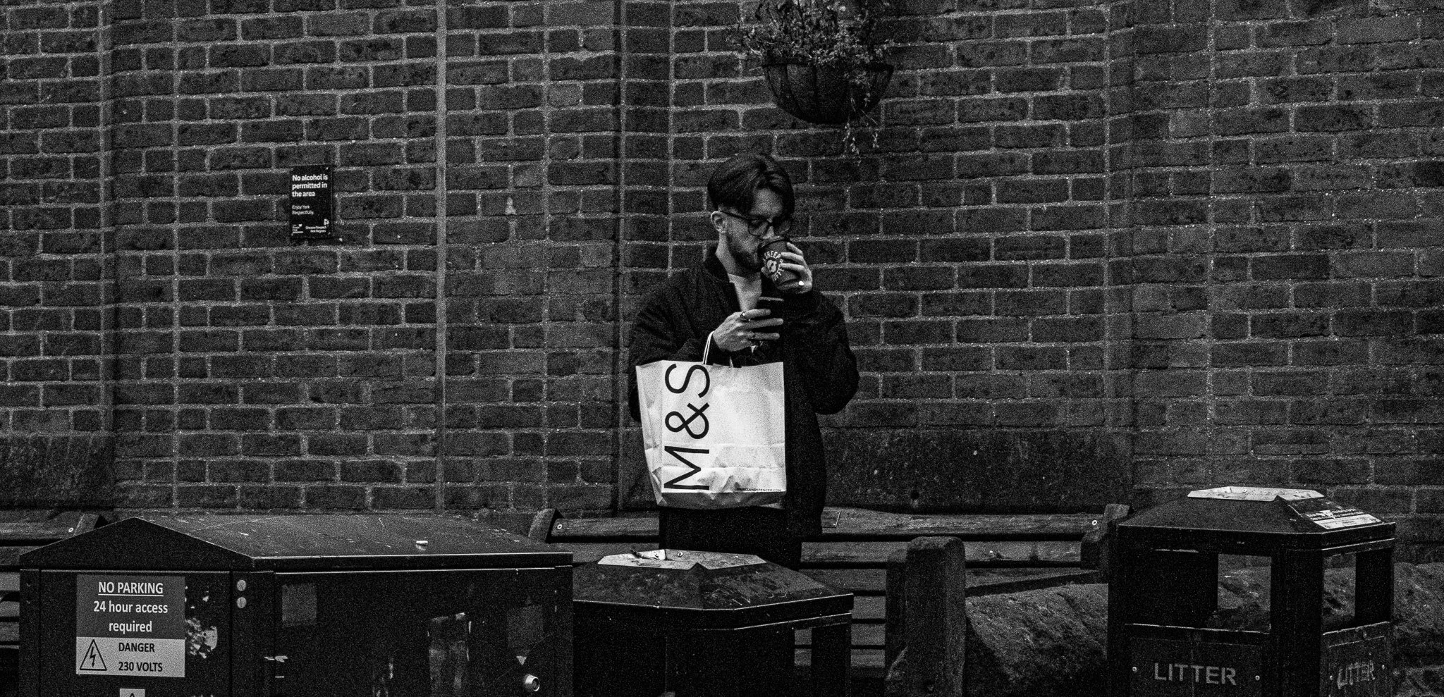 A man with glasses and a beard stands against a brick wall, drinking from a cup and holding a shopping bag, near outdoor trash bins with warning signs.