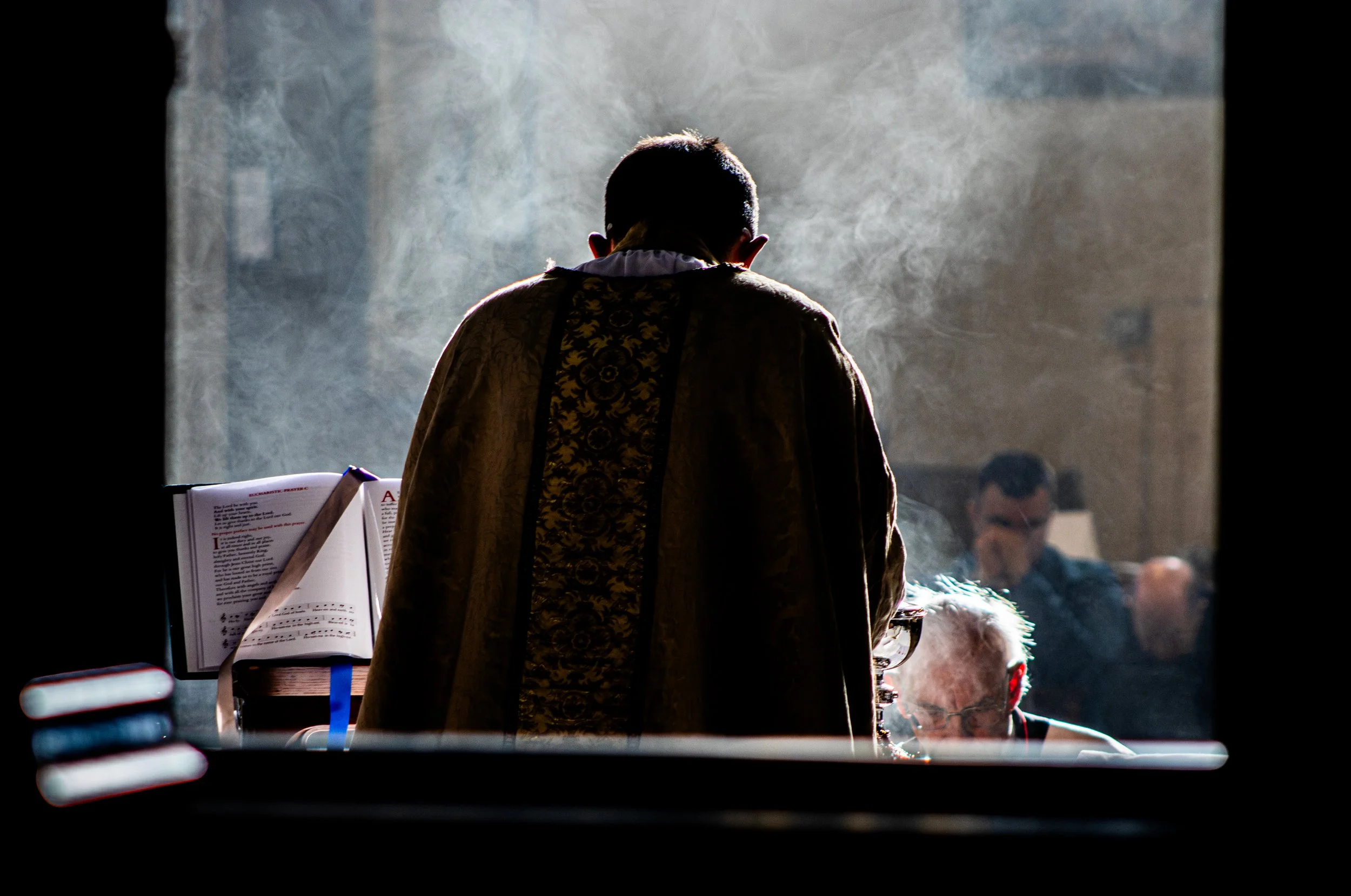 A man with short dark hair dressed in traditional clothing, possibly a jacket with intricate patterns, standing in front of a music stand with sheet music, inside a dimly lit space with a crowd of people in the background.