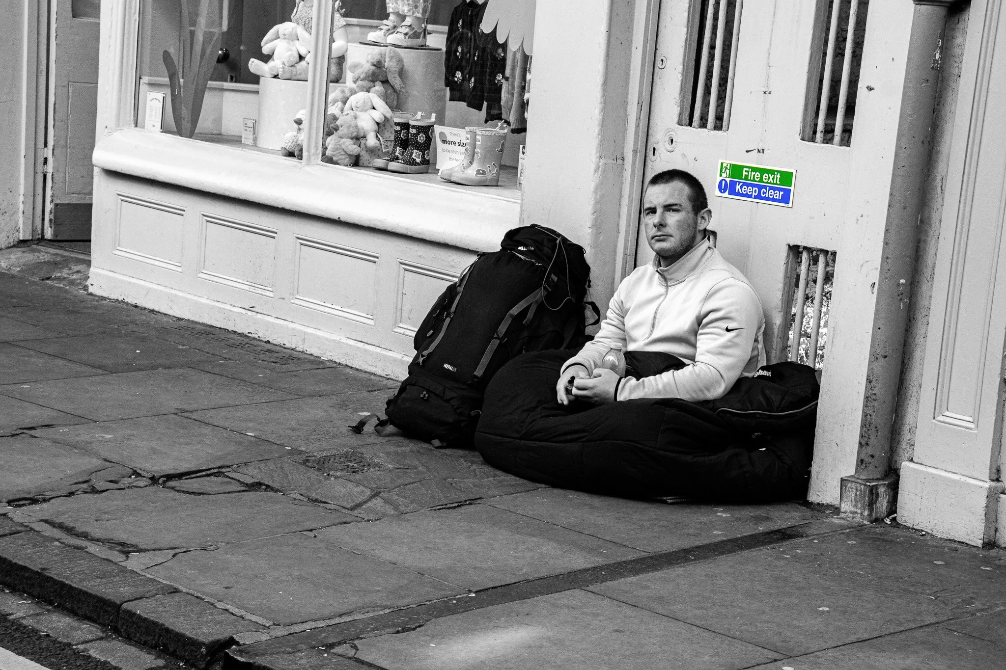 A man in a light-colored jacket sitting on the sidewalk against a building wall, with a large backpack and a black bag beside him. He is holding a water bottle and appears to be resting. Behind him, there is a door with a fire exit and keep clear sig