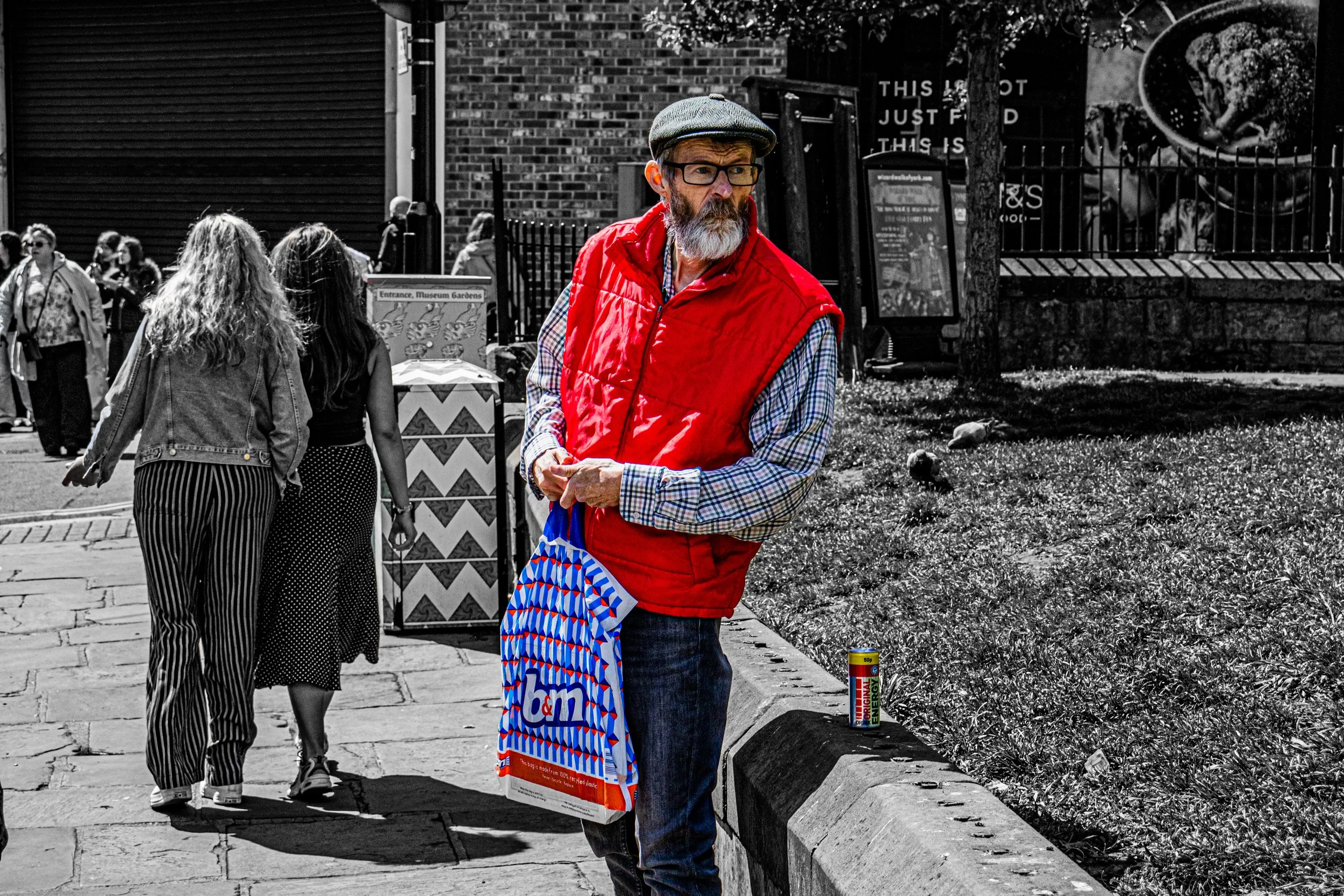A man with a beard and glasses wearing a gray flat cap, red vest, and checkered shirt stands by the sidewalk holding a colorful shopping bag and energy drink can. Behind him, a group of women walk past, and there are signs and graffiti on the brick b