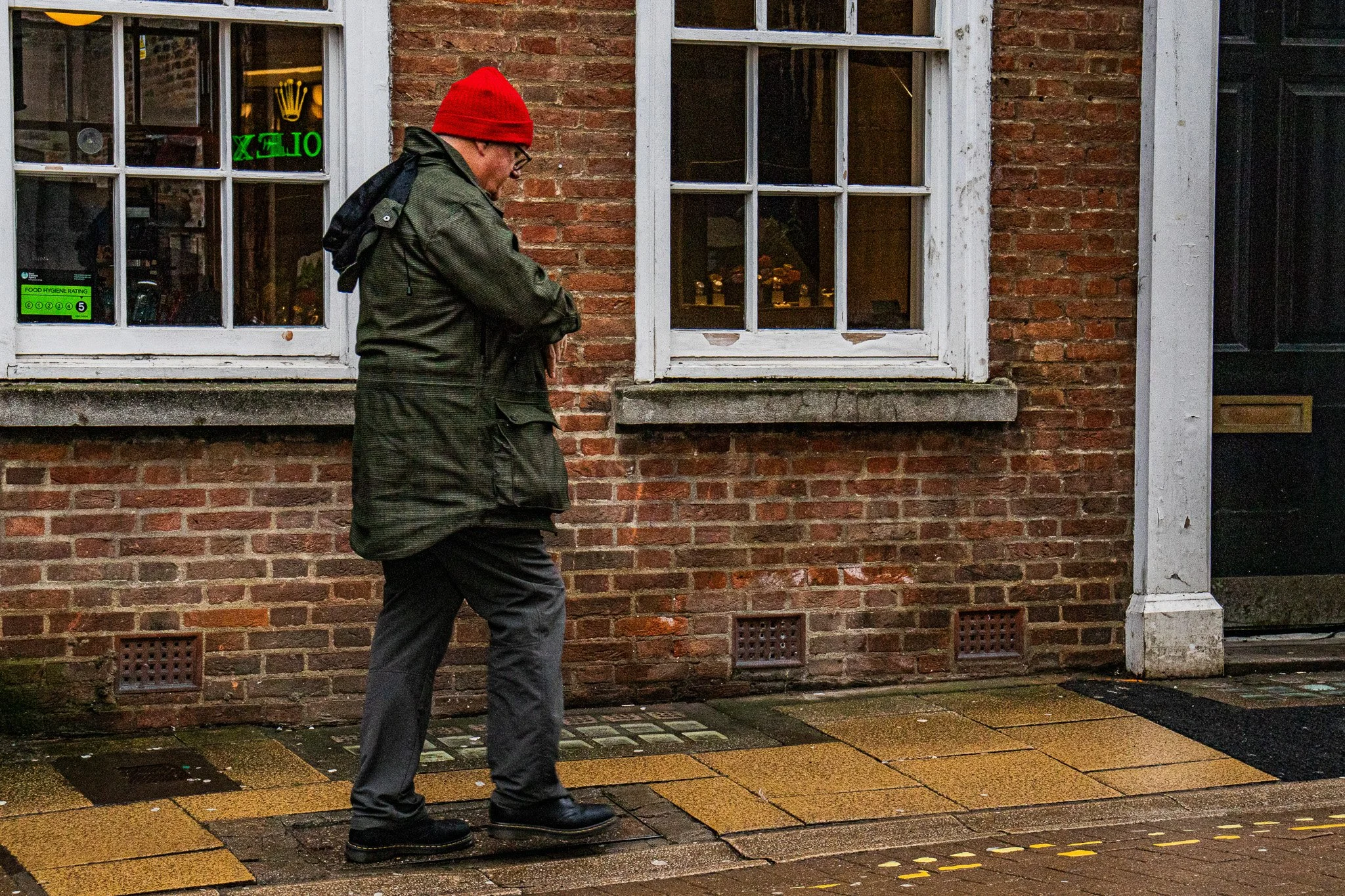Man wearing a red beanie and glasses walking past a brick building with windows on a wet sidewalk in the evening.