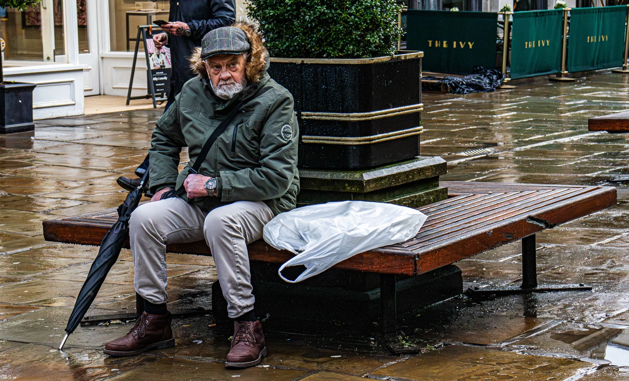 An elderly man with gray hair, beard, and mustache sitting on a wooden bench in front of a building, wearing a green jacket, beige pants, and brown boots, holding a black umbrella and looking at the camera. There is a large white plastic bag on the b
