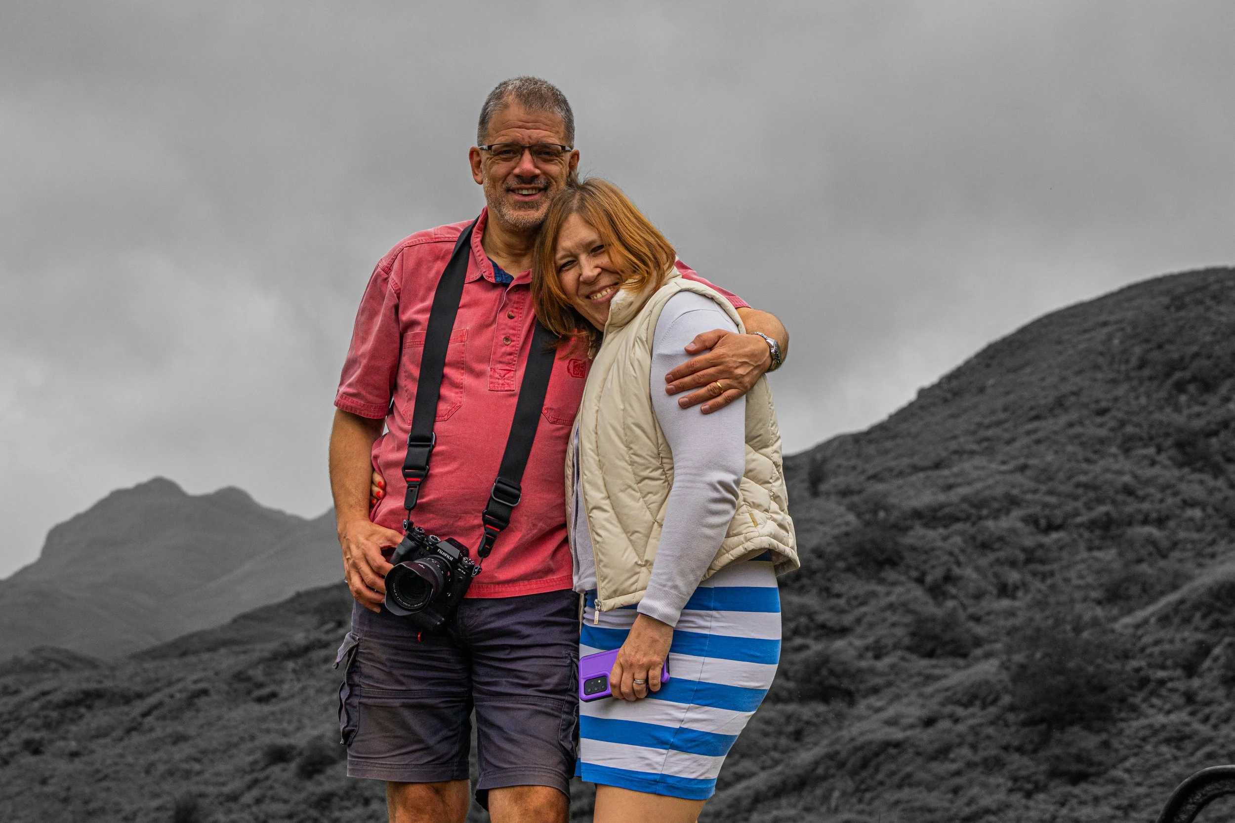 A happy man and woman embrace outdoors against a mountainous, overcast background. The man wears a red shirt and has a camera around his neck, while the woman wears a beige vest and a striped dress, holding a purple phone.