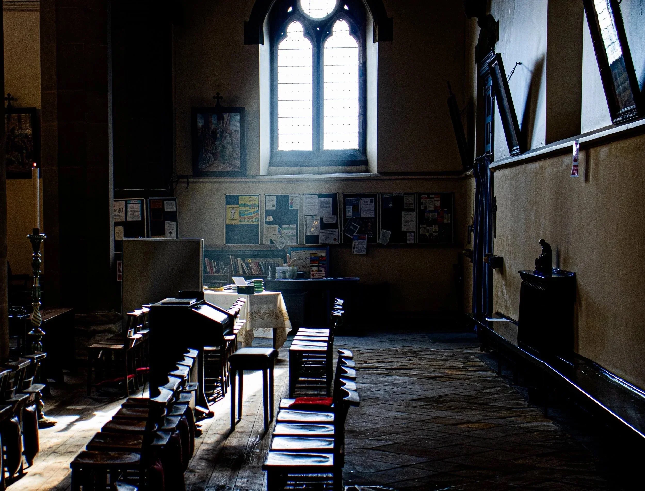 Interior of a church or chapel with wooden chairs lined up along the aisle, lit by natural light coming through a large stained-glass window at the back. The scene appears dark with shadows, and religious artwork is displayed on the walls.