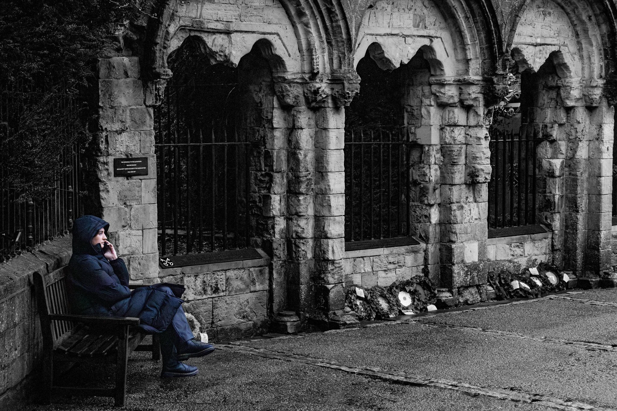 A woman sitting on a bench, talking on her phone, dressed in a dark jacket and pants, next to an old stone wall with arches and a wreath with flowers at its base.