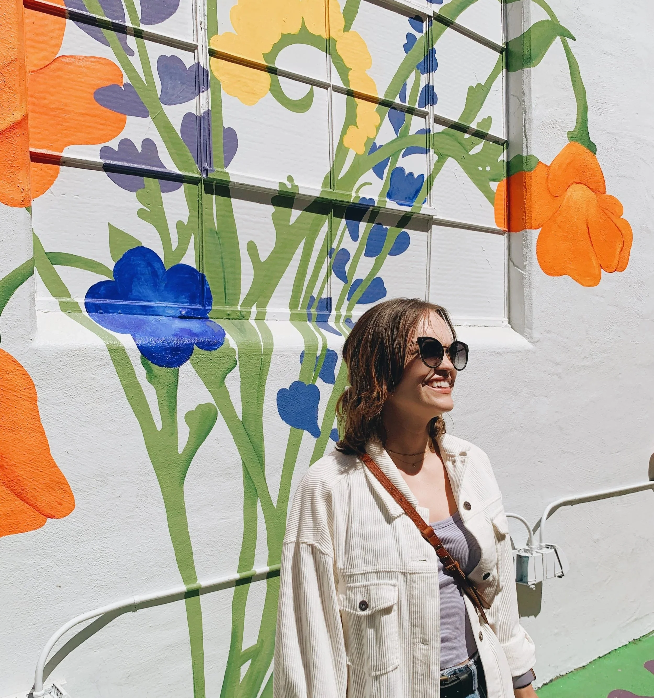 Kate, a woman with shoulder-length brown hair and sunglasses, smiles while standing in front of a colorful floral mural on a white wall.