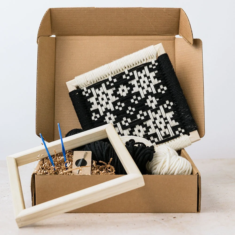 Open cardboard box containing black and white woven wall hanging, balls of black and white yarn, and a small framed craft project with two blue craft sticks.
