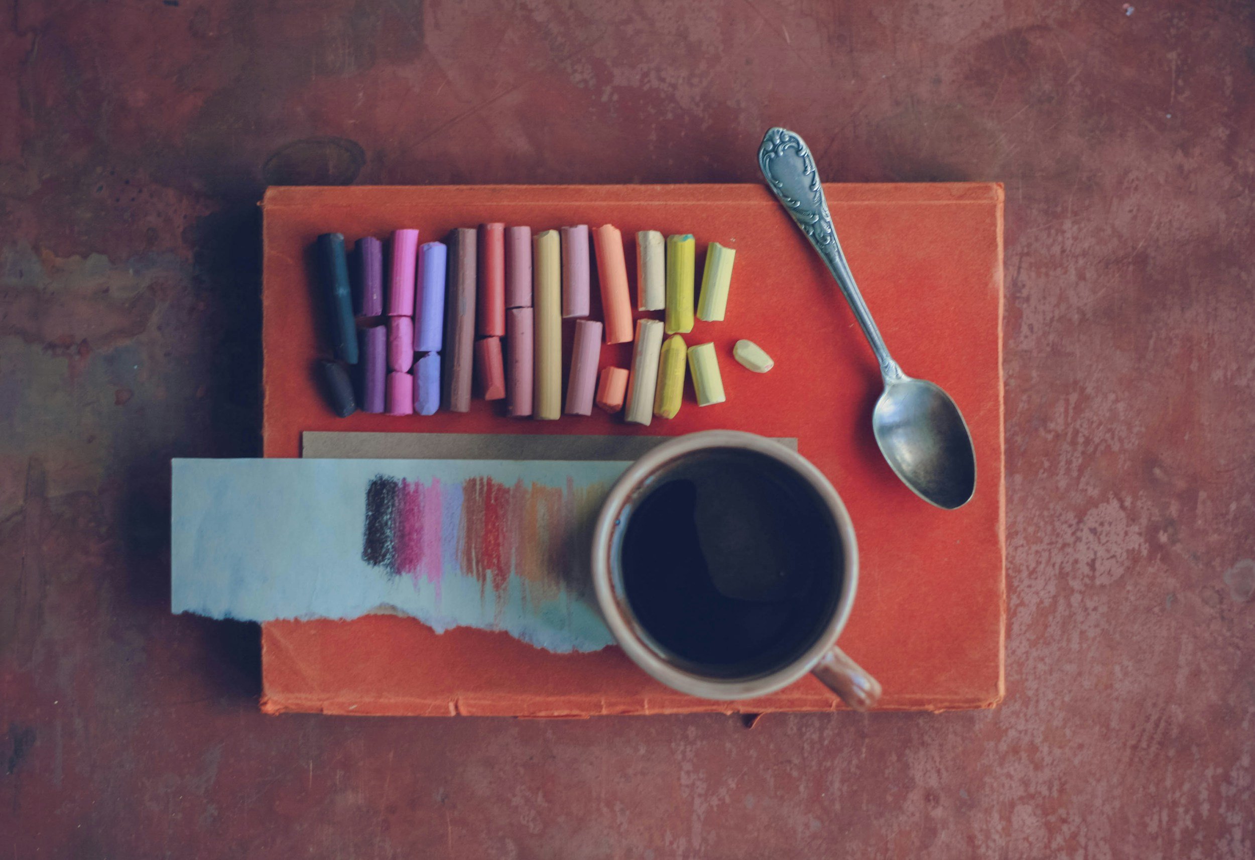 A cup of black coffee on a saucer, a colorful assortment of chalk pieces on an orange cloth, a silver spoon, and a small painting with colorful strokes, all placed on a weathered wooden surface.