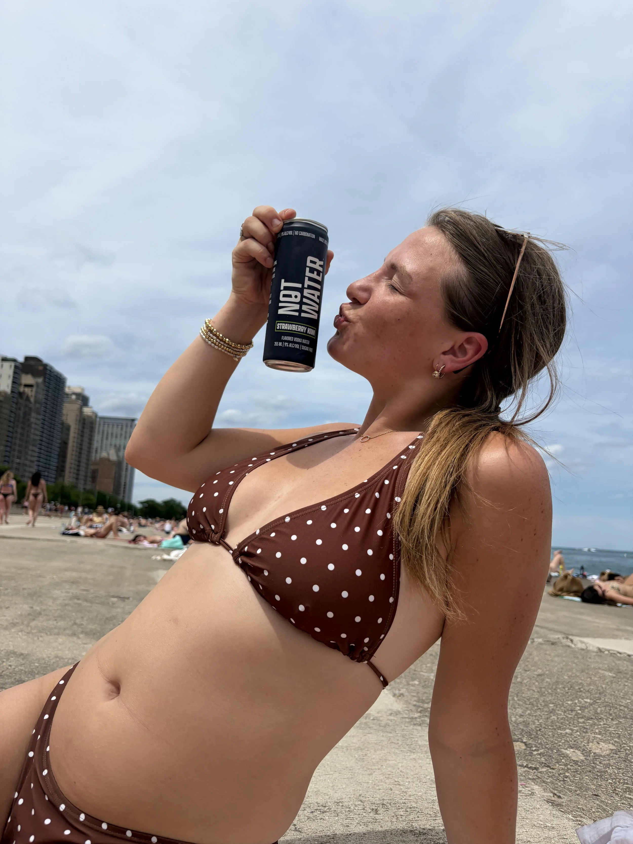 A woman in a brown polka dot bikini sitting on a beach holding a 'Not Water' flavored vodka can near her face, with Chicago skyline and other beachgoers in the background on a cloudy day.