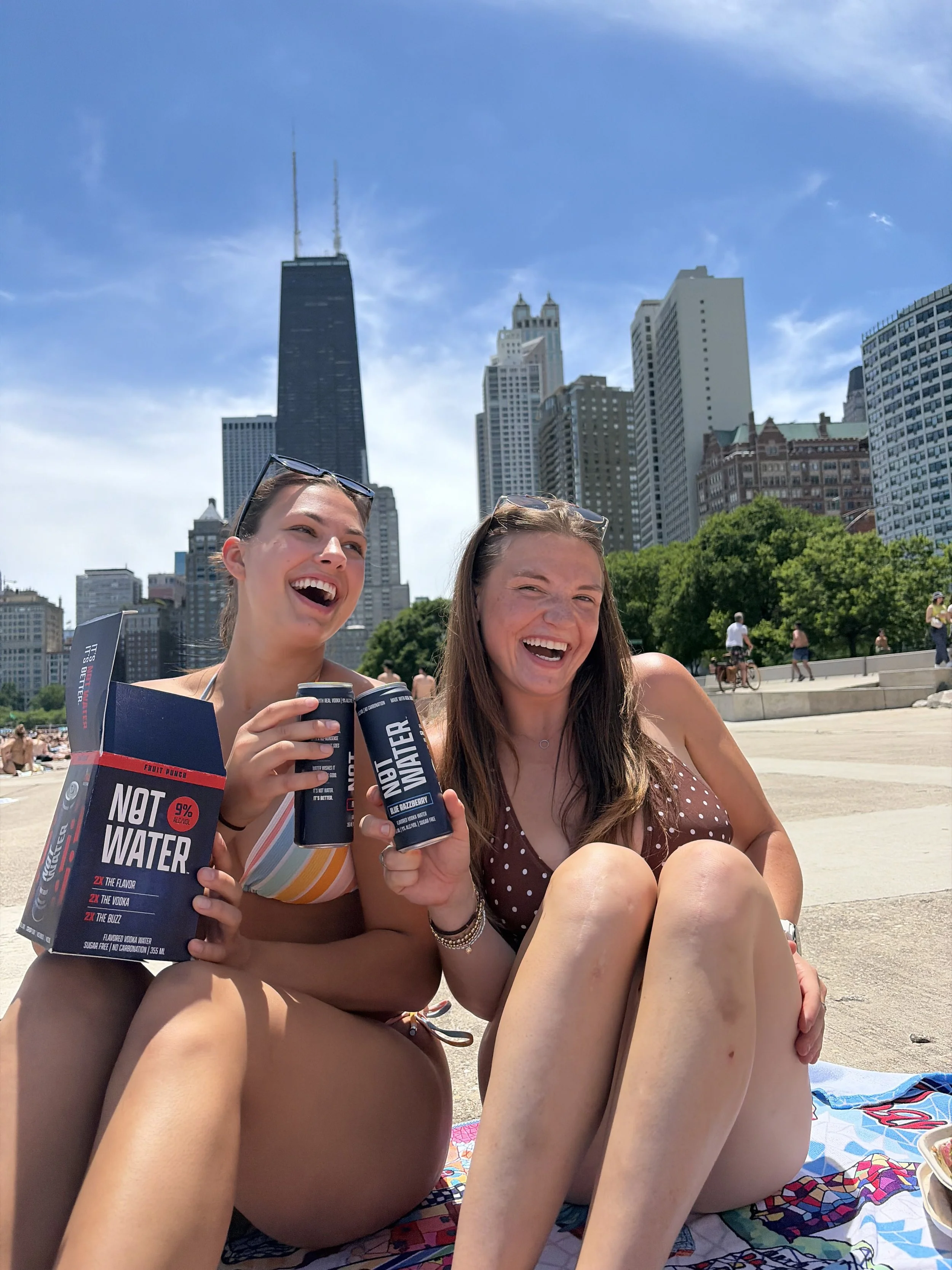 Two women sit on a colorful towel at the beach, smiling and holding cans of NOT WATER, a 9% ABV alcoholic beverage with no carbonation, zero sugar, and gluten free ingredients. A city skyline featuring the Willis Tower and Chicago buildings appear.