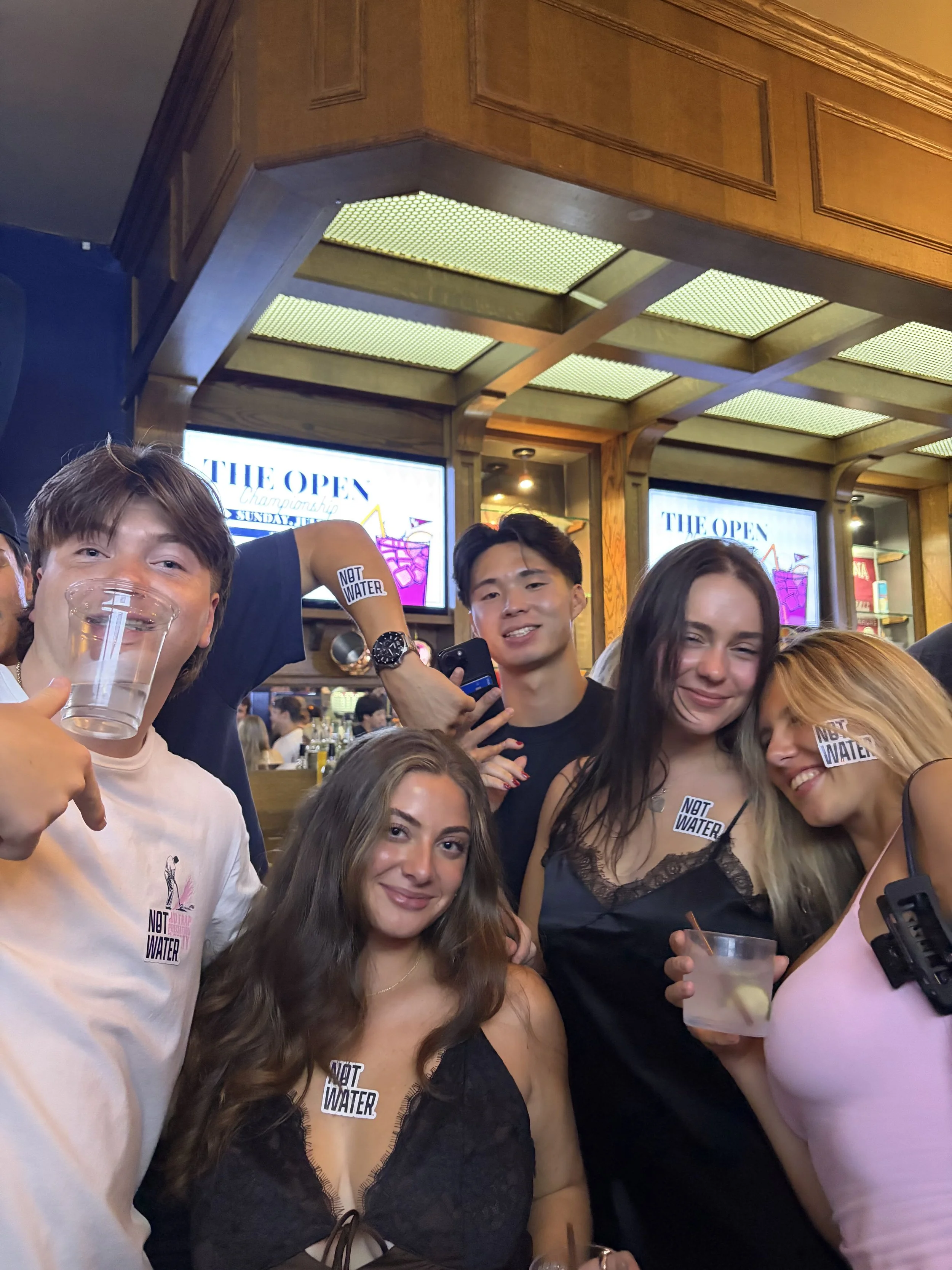 A group of people gathers at a bar or restaurant, smiling and holding cans of NOT WATER, a 9% ABV alcoholic beverage with no carbonation, zero sugar, and gluten free ingredients. Some people are wearing “NOT WATER” stickers.