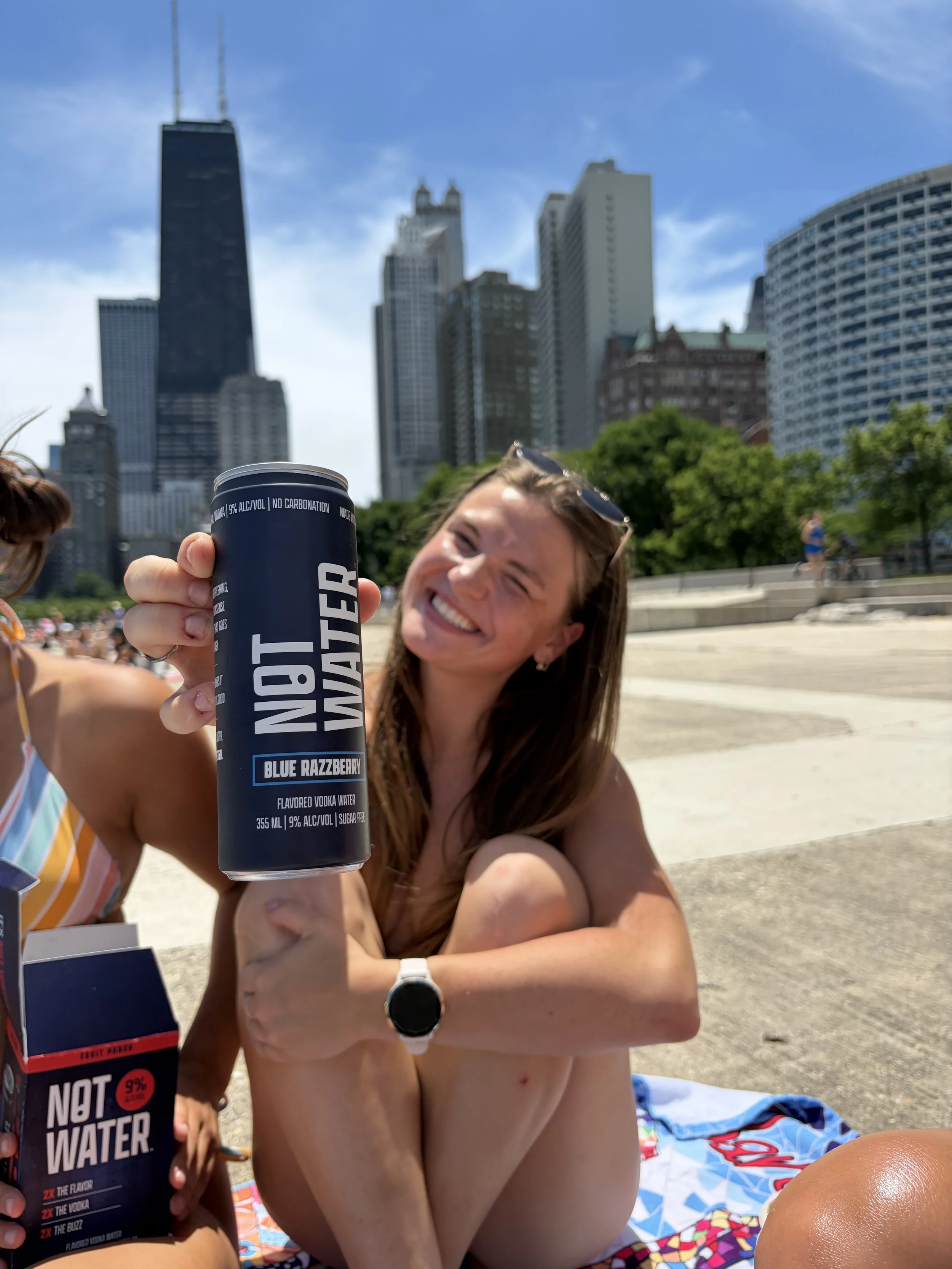 A smiling woman with long hair and sunglasses on her head is sitting on a colorful towel outdoors in a city park, holding a can of NOT WATER Blue Razzberry flavored vodka water. The background features tall skyscrapers and green trees.