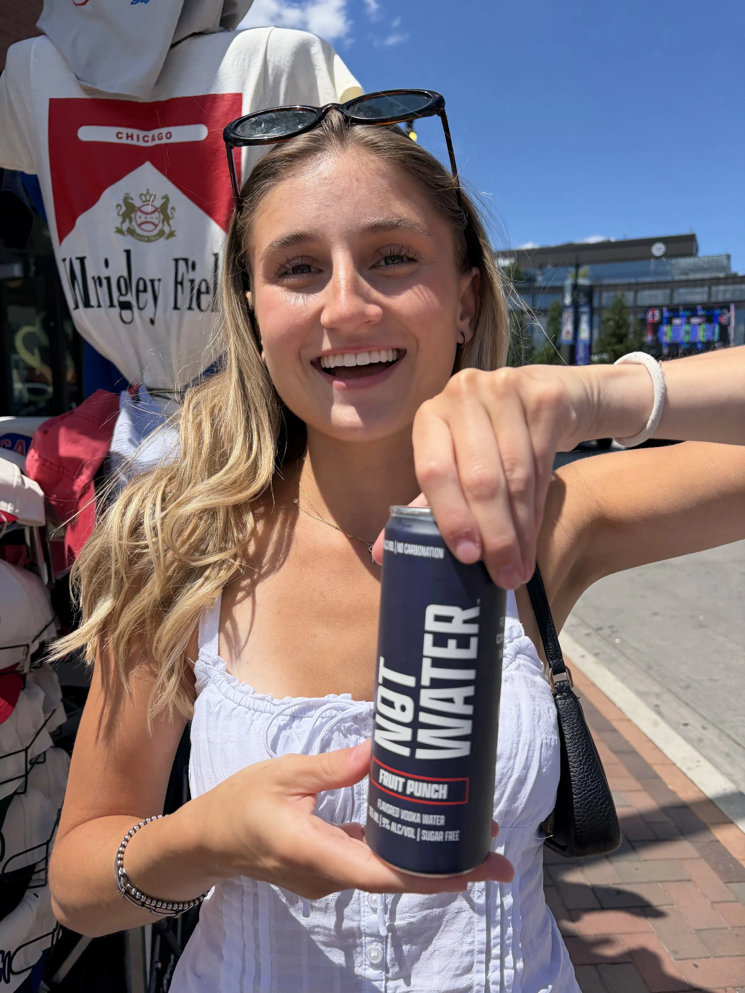 A young woman with long blonde hair, sunglasses on her head, smiling while holding a bottle of fruit punch Not Water