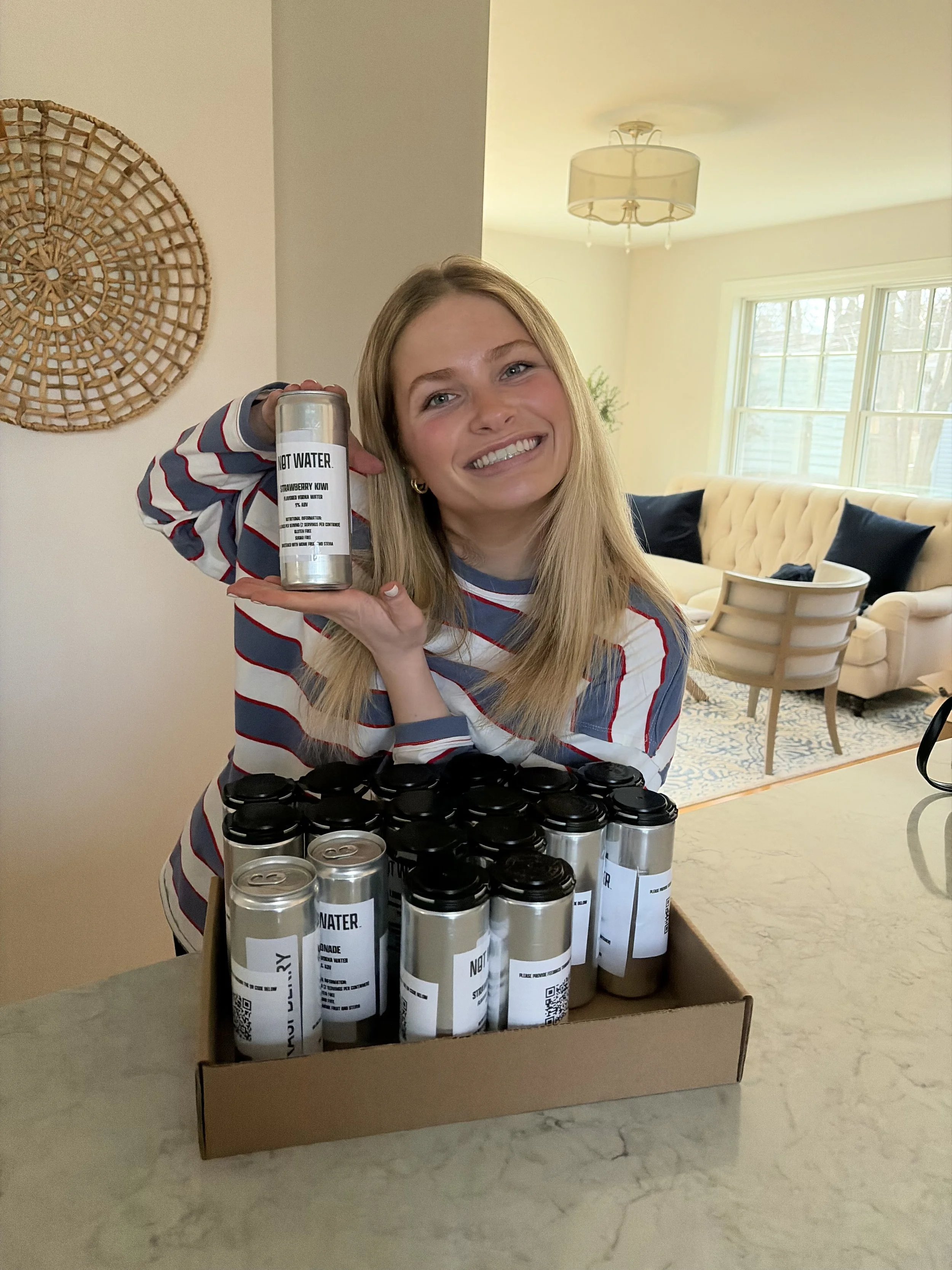 A smiling woman holds a canned alcoholic beverage labeled “NOT WATER” beside a cardboard box filled with similar cans. The 9% ABV, no carbonation, zero sugar, gluten free beverage is featured in a bright living room with large windows, a beige couch,