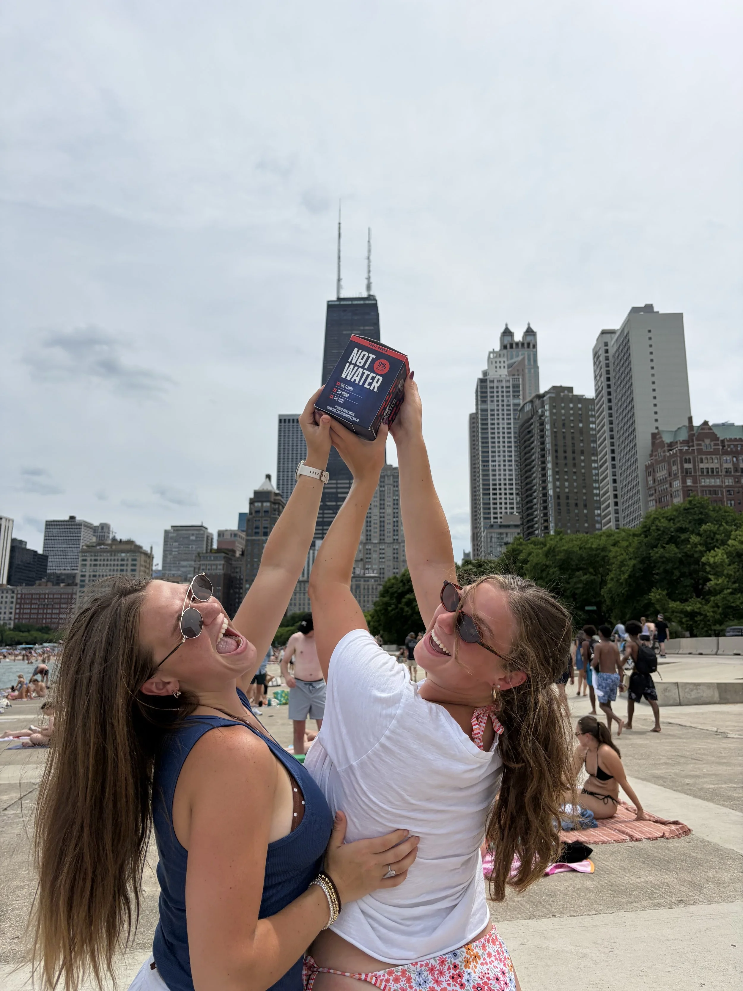 Two women smiling and laughing, holding a small box with the words "NOT WATER" at a park with a city skyline and Chicago, Illinois in the background during daytime.