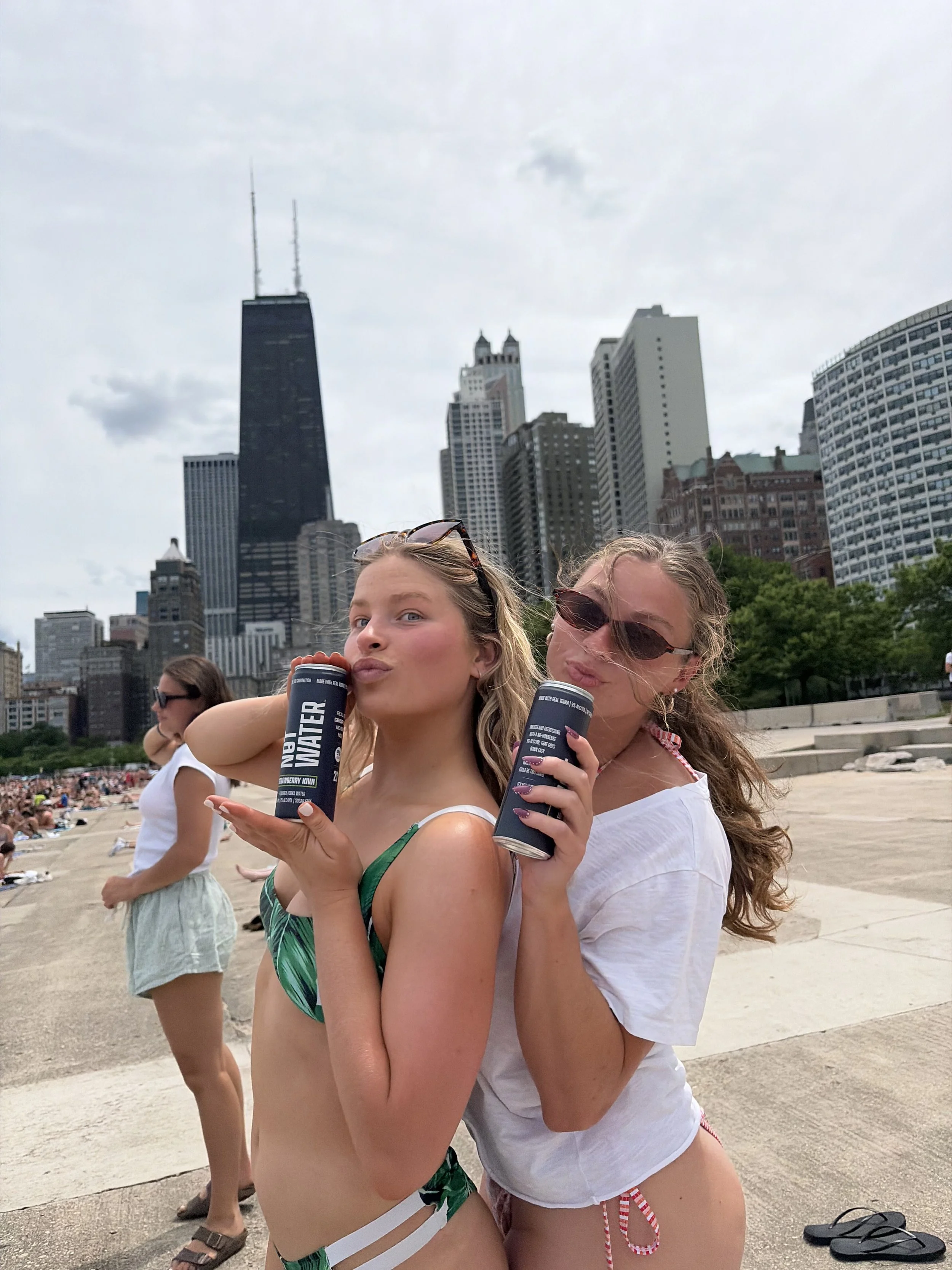 Two young women stand together at a crowded outdoor location along Lake Michigan in downtown Chicago, smiling and holding cans of NOT WATER, a 9% ABV alcoholic beverage with no carbonation, zero sugar, and gluten free ingredients.