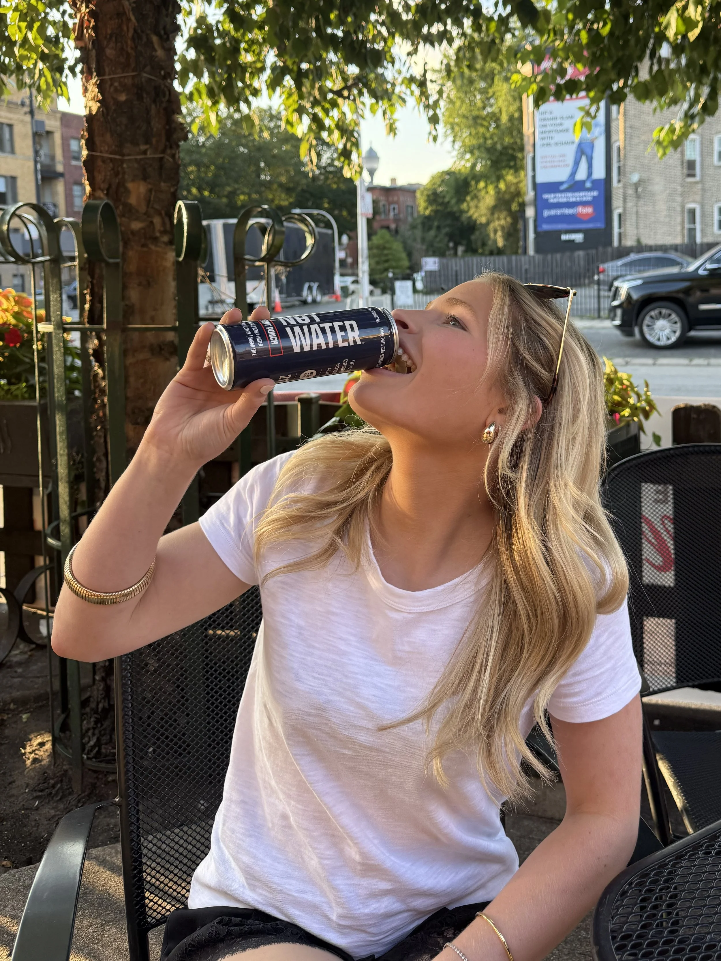 A young woman with blonde hair wearing a white t-shirt and earrings, drinking 9% ABV vodka 'Not Water'