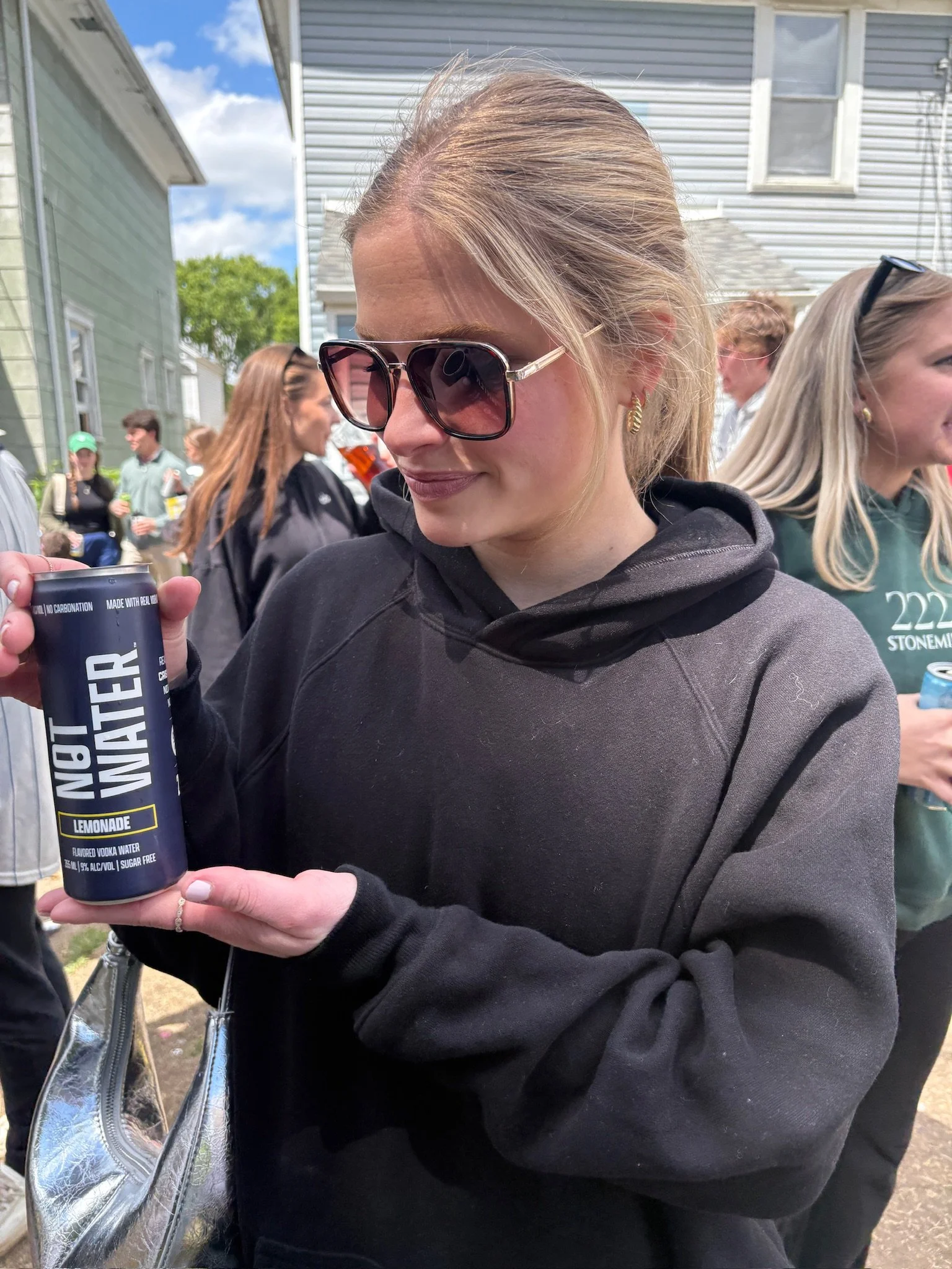 A blonde woman wearing large sunglasses and a black hoodie holds a can of NOT WATER, a 9% ABV alcoholic beverage with no carbonation, zero sugar, and gluten free ingredients. She is outdoors with a house and other people visible in the background.