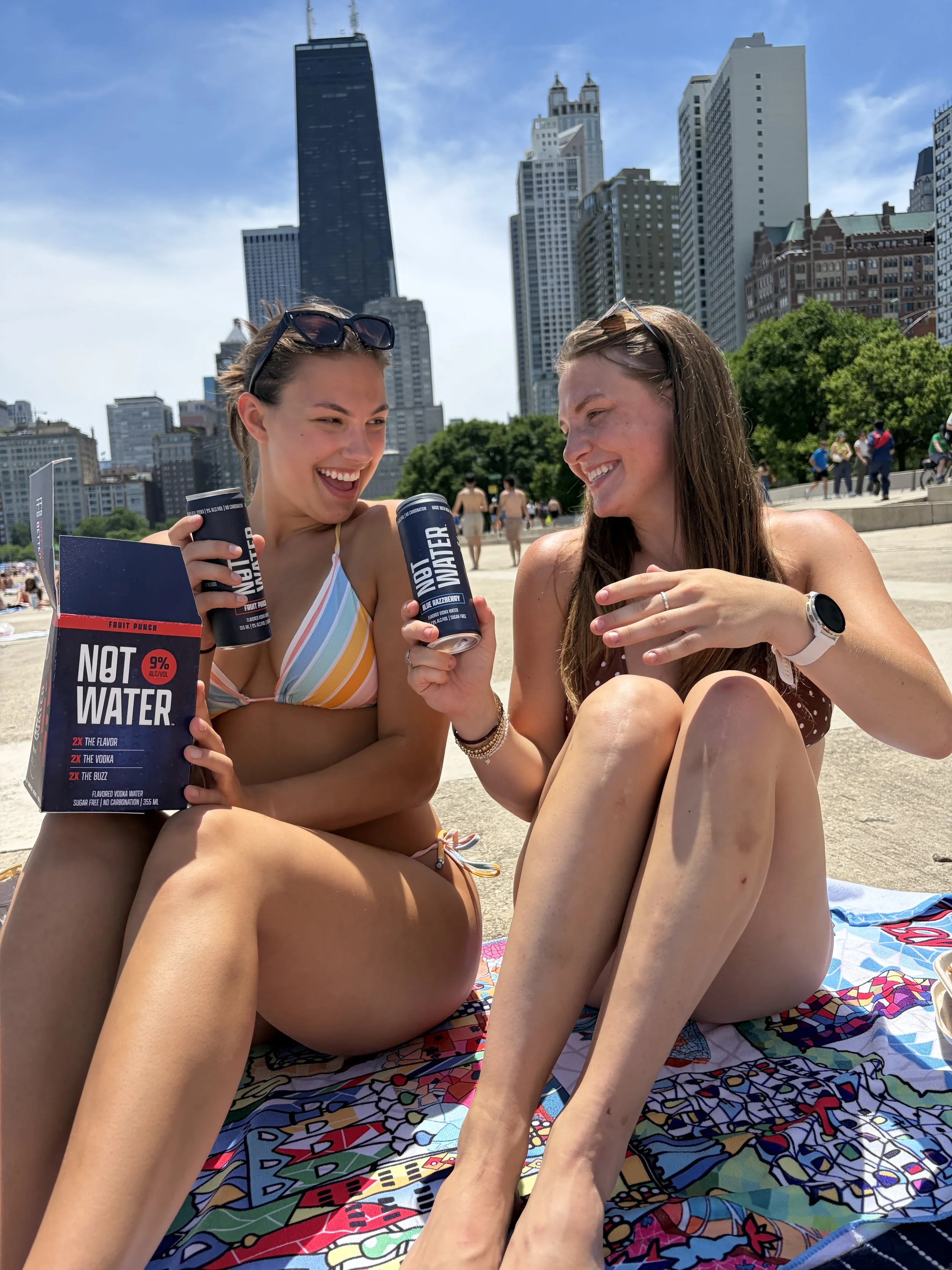 Two young women in swimsuits sitting on a colorful beach towel at the beach, holding cans of Not Water, smiling and talking, with Chicago, Illinois city skyline in the background on a sunny day.