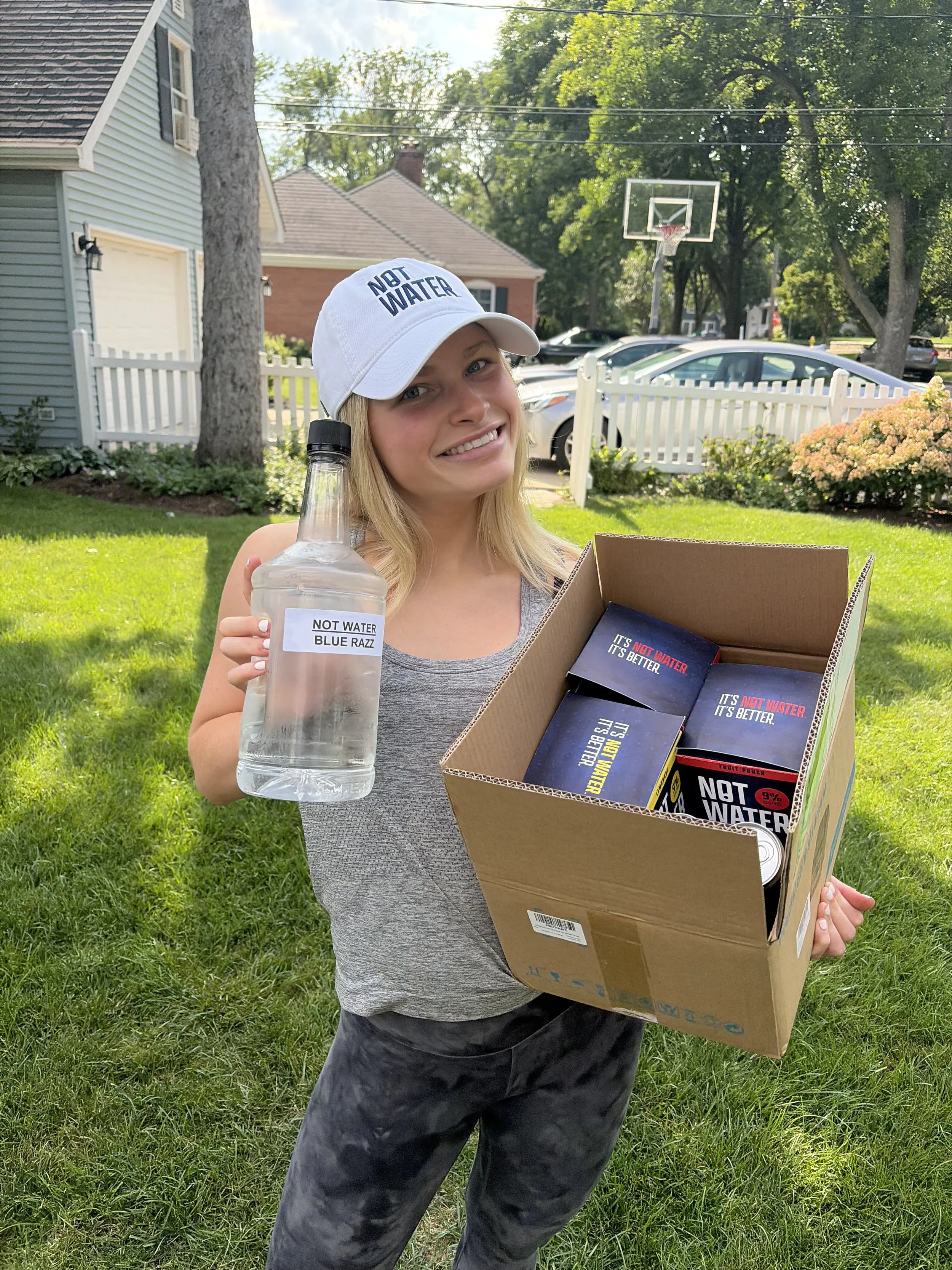 A woman outdoors holding an alcoholic beverage with 9% ABV, no carbonation, zero sugar, and gluten free ingredients. She is wearing a white cap with blue text and a gray tank top. The background includes a house, fence, trees, parked cars, and a bask