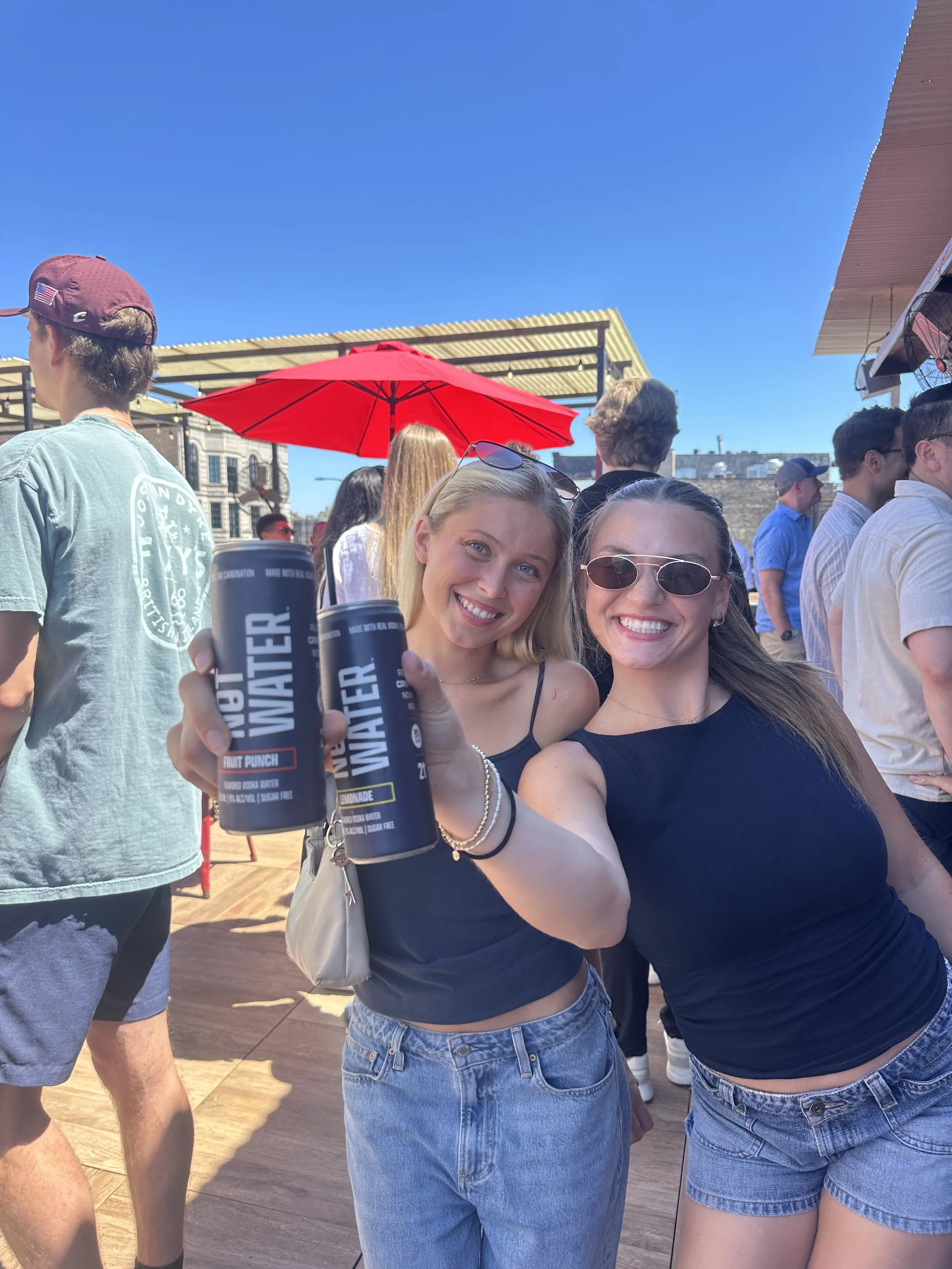 Two smiling women in tank tops and denim shorts hold cans of NOT WATER, a 9% ABV alcoholic beverage with no carbonation, zero sugar, and gluten free ingredients, at an outdoor event.