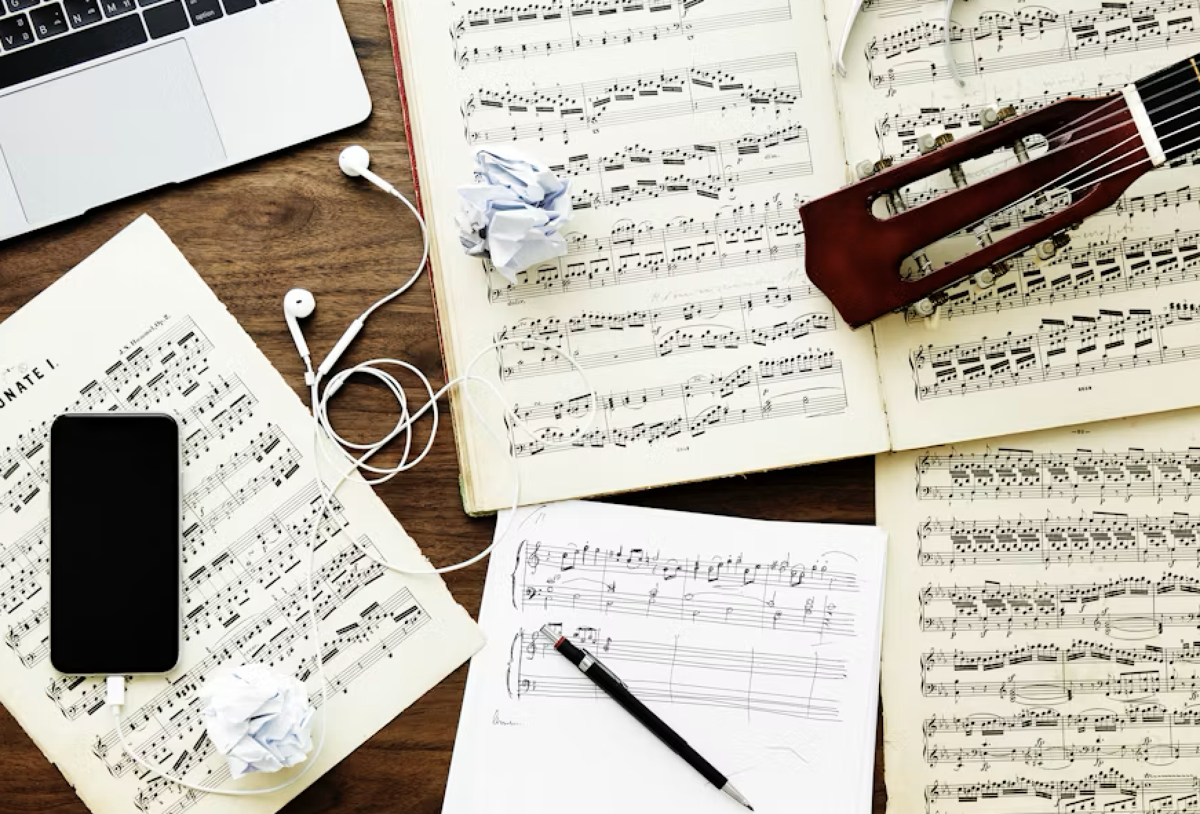 Music sheets, a pen, crumpled paper, a smartphone, earphones, a laptop, and a violin on a wooden table.