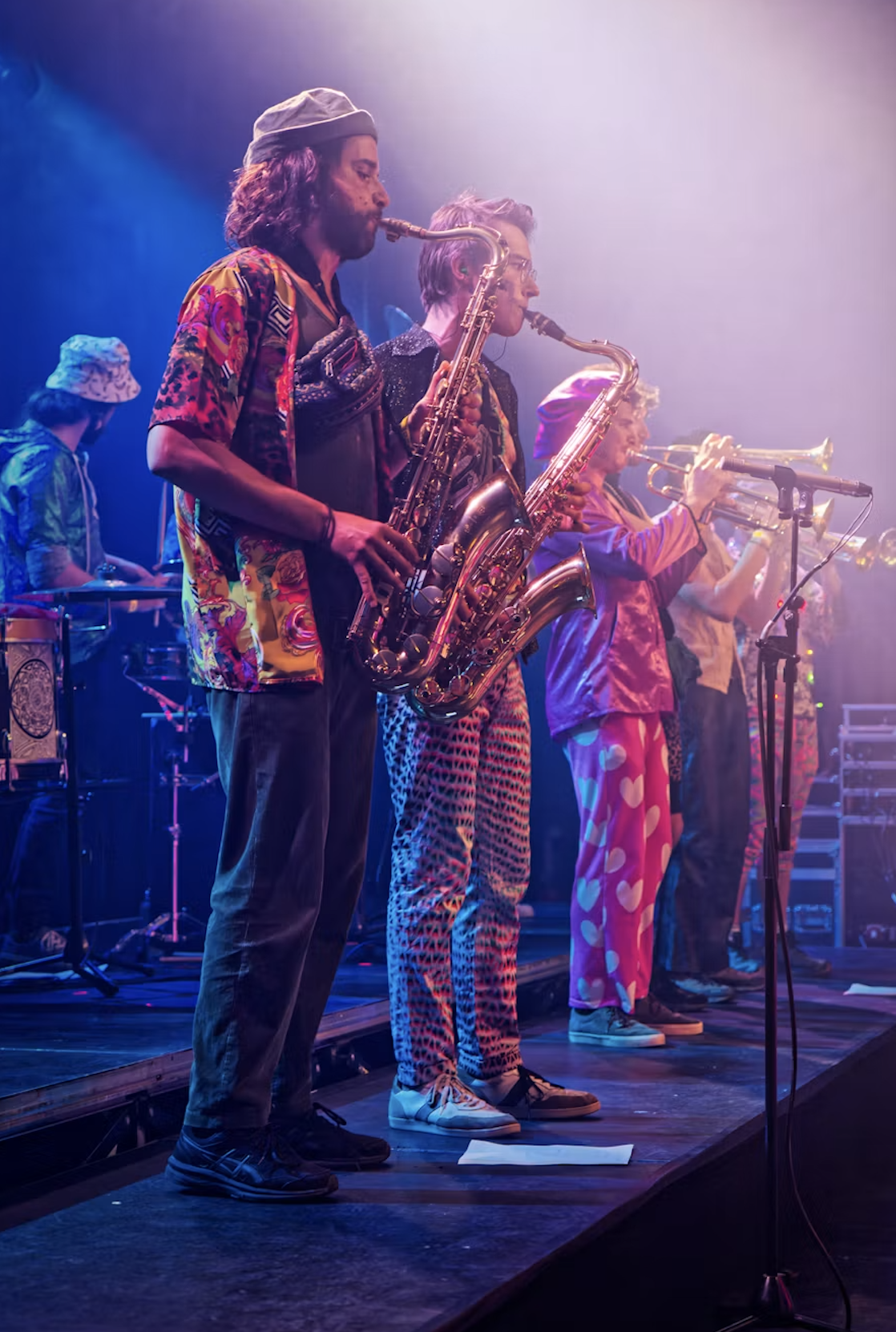 Band performing on stage with multiple musicians playing saxophones and trumpets, wearing colorful and patterned clothing, under stage lights.