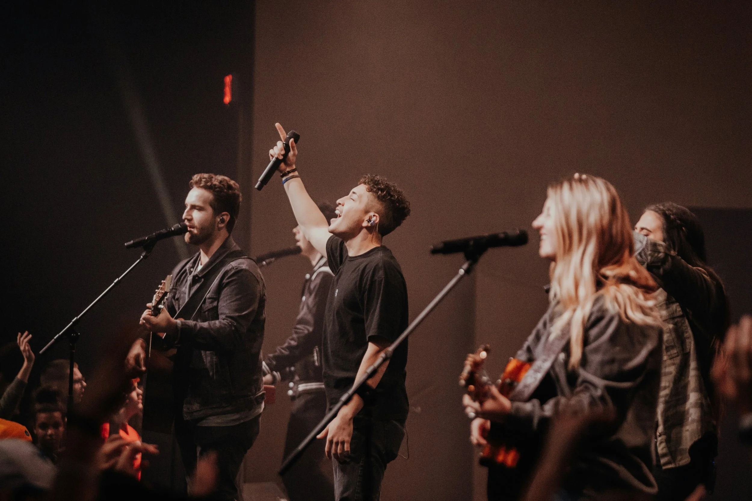 A group of people, including young men and women, are performing on stage with microphones and guitars, with an audience visible in the foreground.