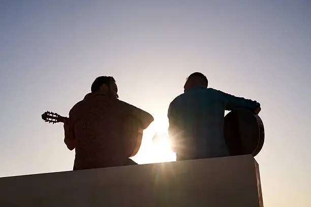 Two people playing guitars outdoors at sunset, seen from behind.