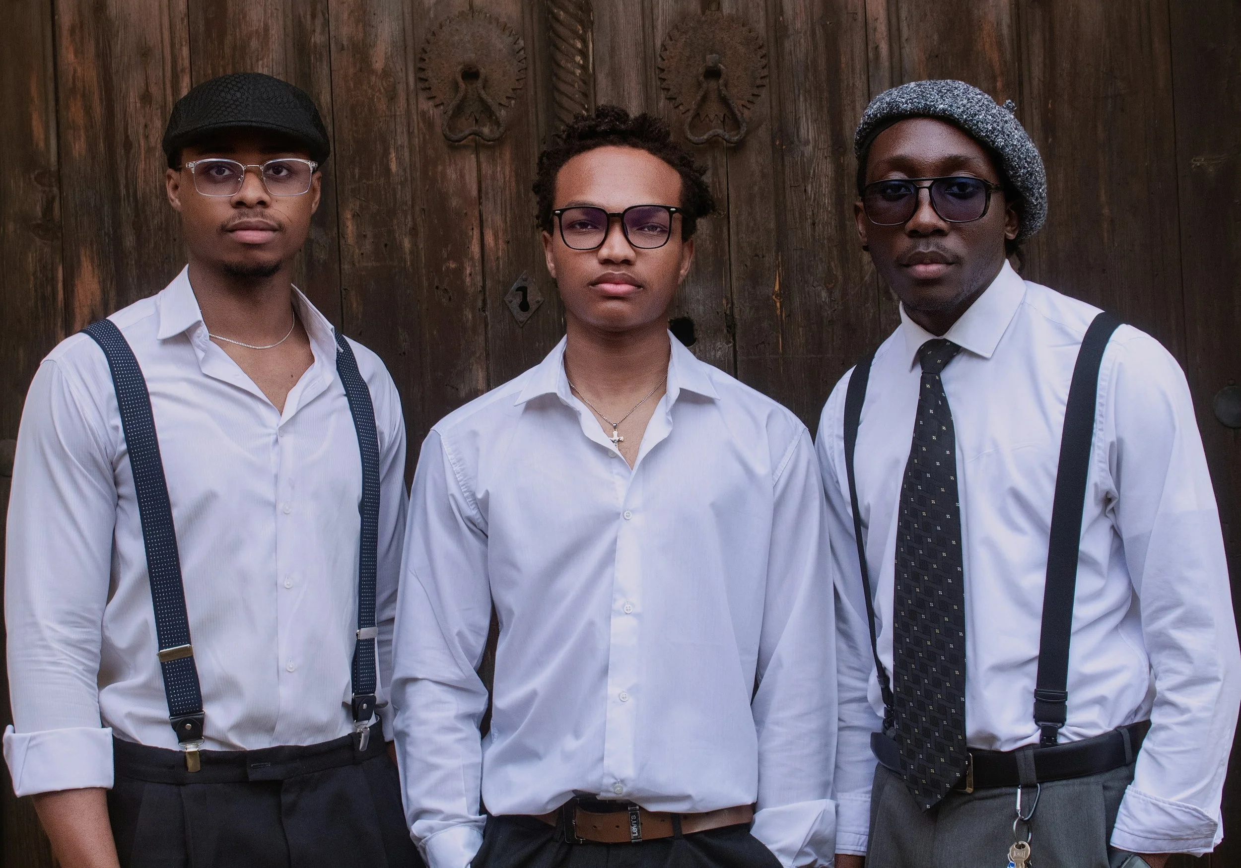 Three young adults posing in front of a wooden door with metal decorative hardware. They are dressed in vintage style with white shirts, suspenders, and accessories like glasses and hats.
