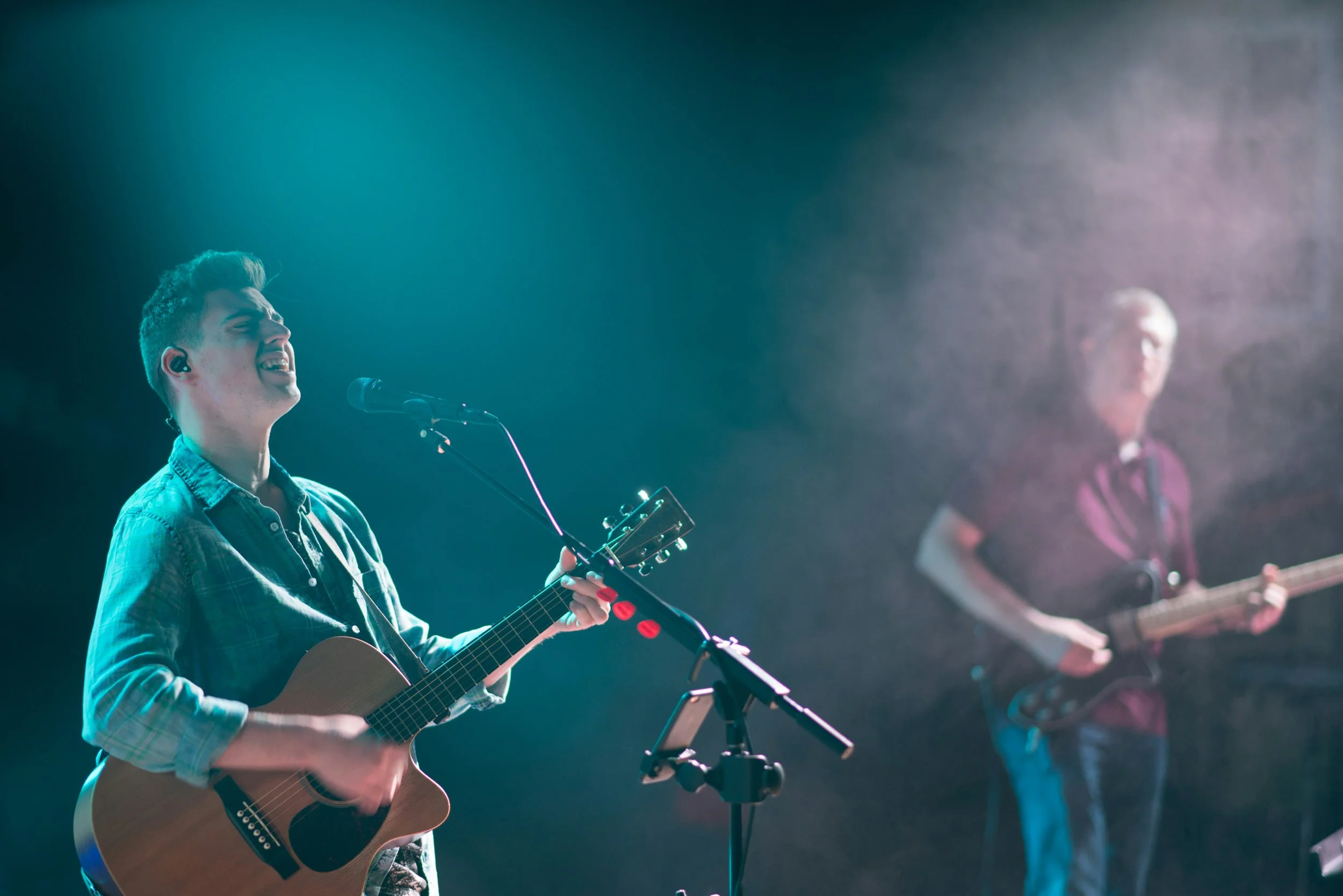 A young male musician with short hair singing into a microphone while playing an acoustic guitar during a live performance, with another musician playing guitar in the background, both under stage lighting.