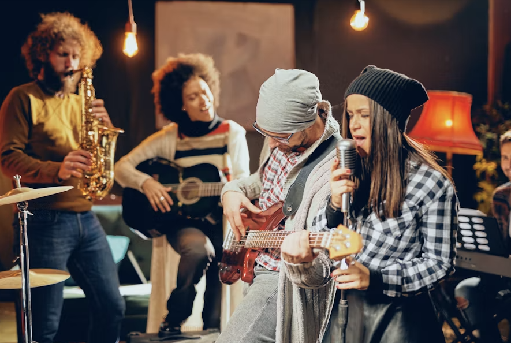 A group of musicians performing together in a cozy indoor setting. One woman is singing into a microphone while playing an acoustic guitar, another woman in the background is playing a different guitar, a man is playing the keyboard, and another person is playing the saxophone. Warm lighting and homey decor create an intimate atmosphere.