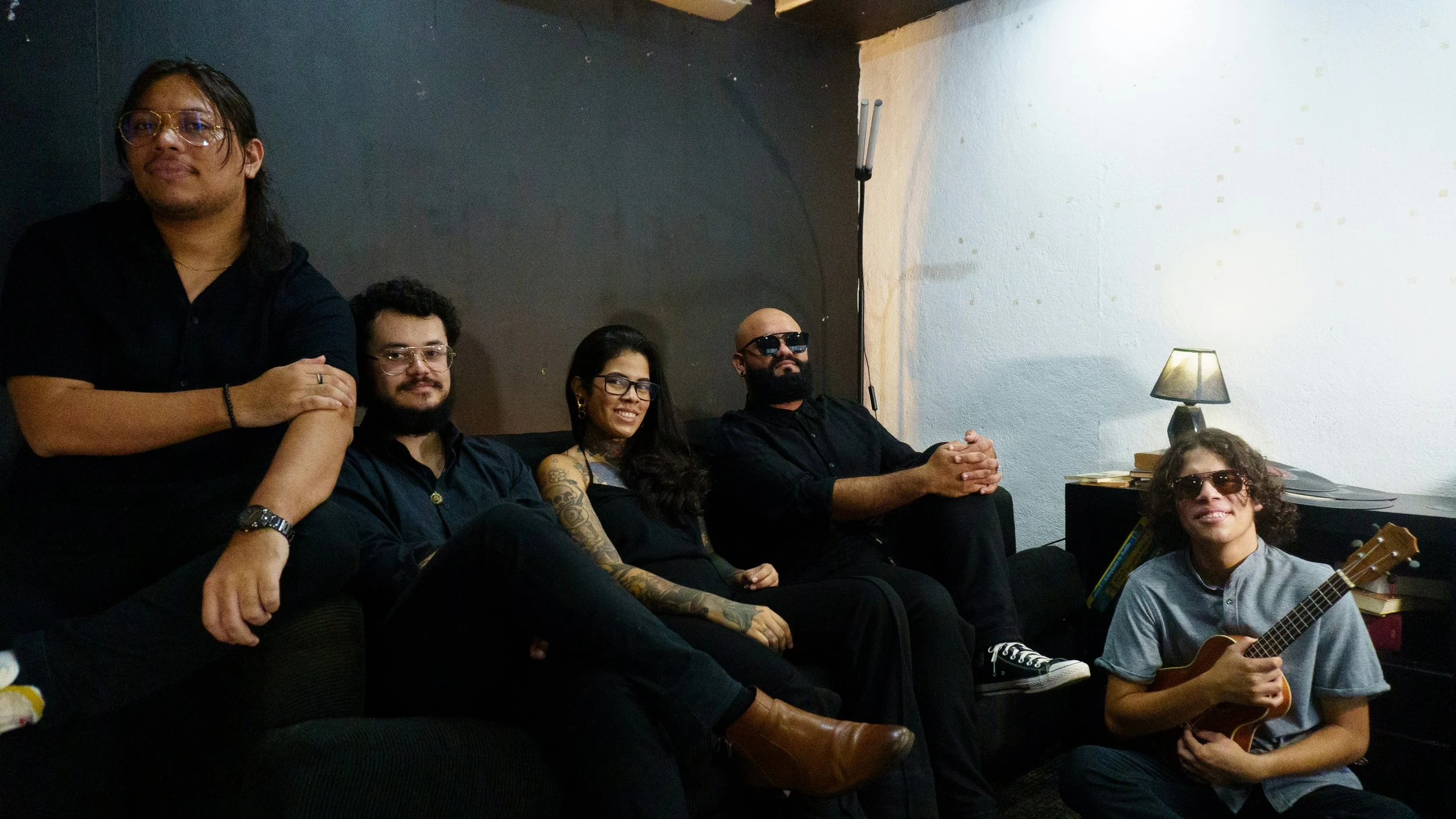A group of six people sitting and standing on a couch and chair against a black and white wall, with one person holding a guitar and another wearing sunglasses, in a cozy indoor setting.