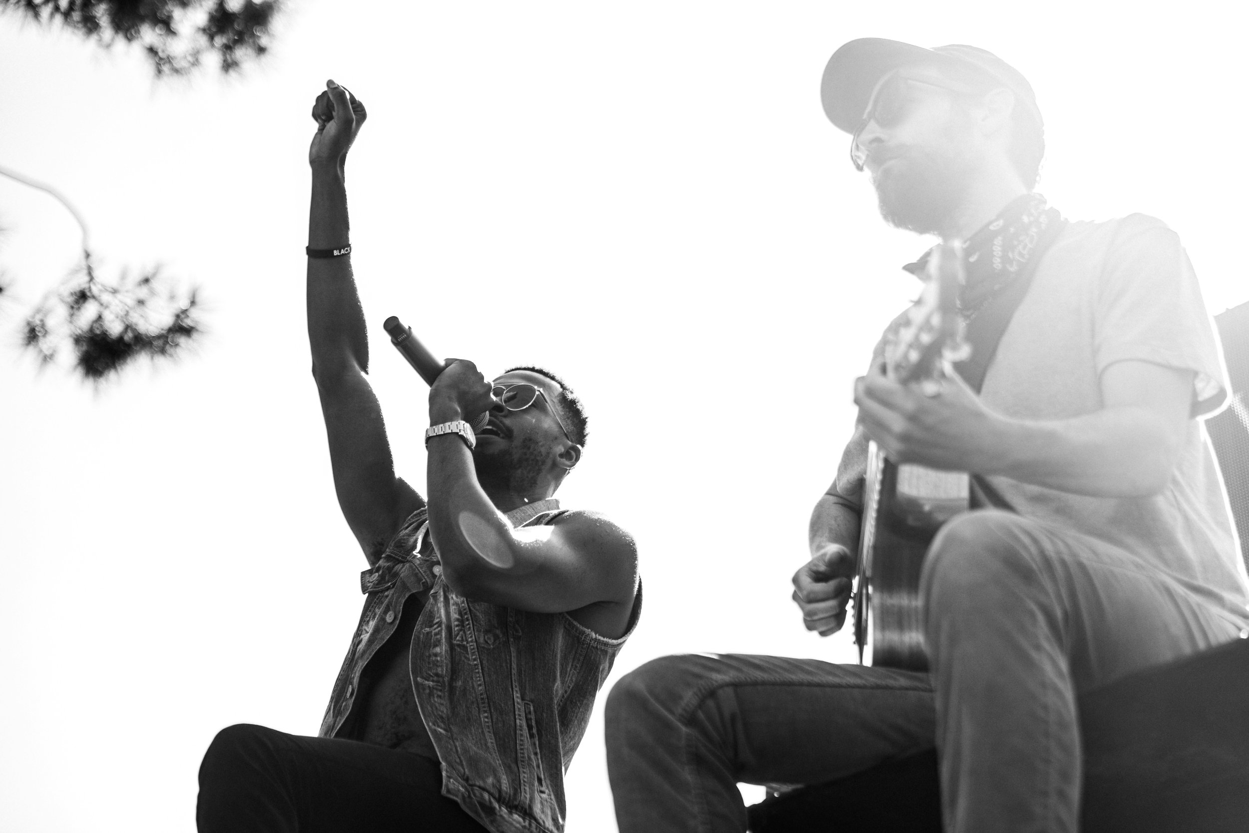 A person with sunglasses and a sleeveless shirt singing into a microphone with their arm raised, and a man playing an acoustic guitar looking at them in an outdoor setting, with trees in the background.