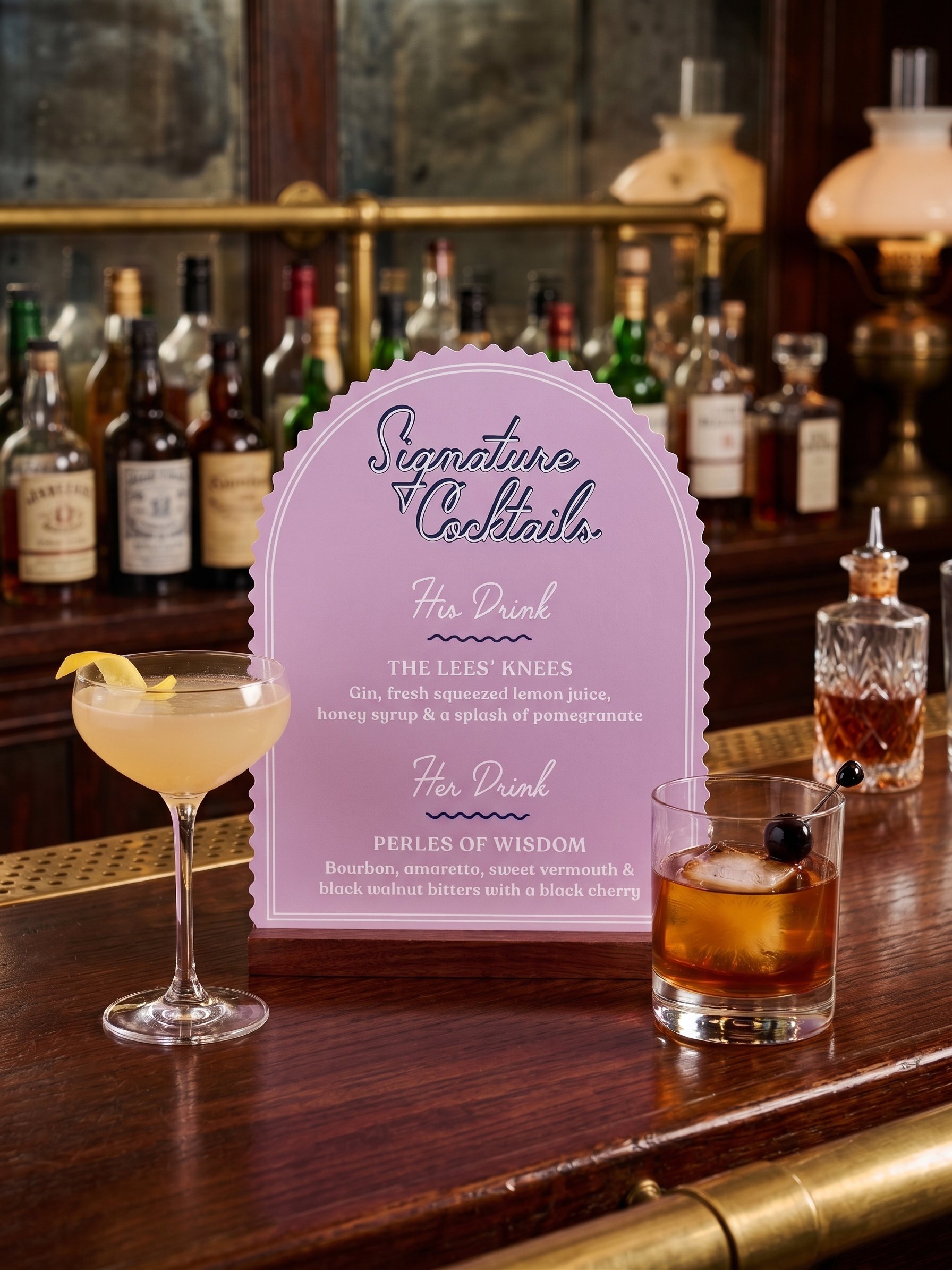 Two cocktails on a wooden bar: one in a coupe glass with a lemon twist and the other in a rocks glass with cherries and ice. A purple sign behind the drinks reads 'Signature Cocktails, Her Drink, Perles of Wisdom' and describes the ingredients of each drink.